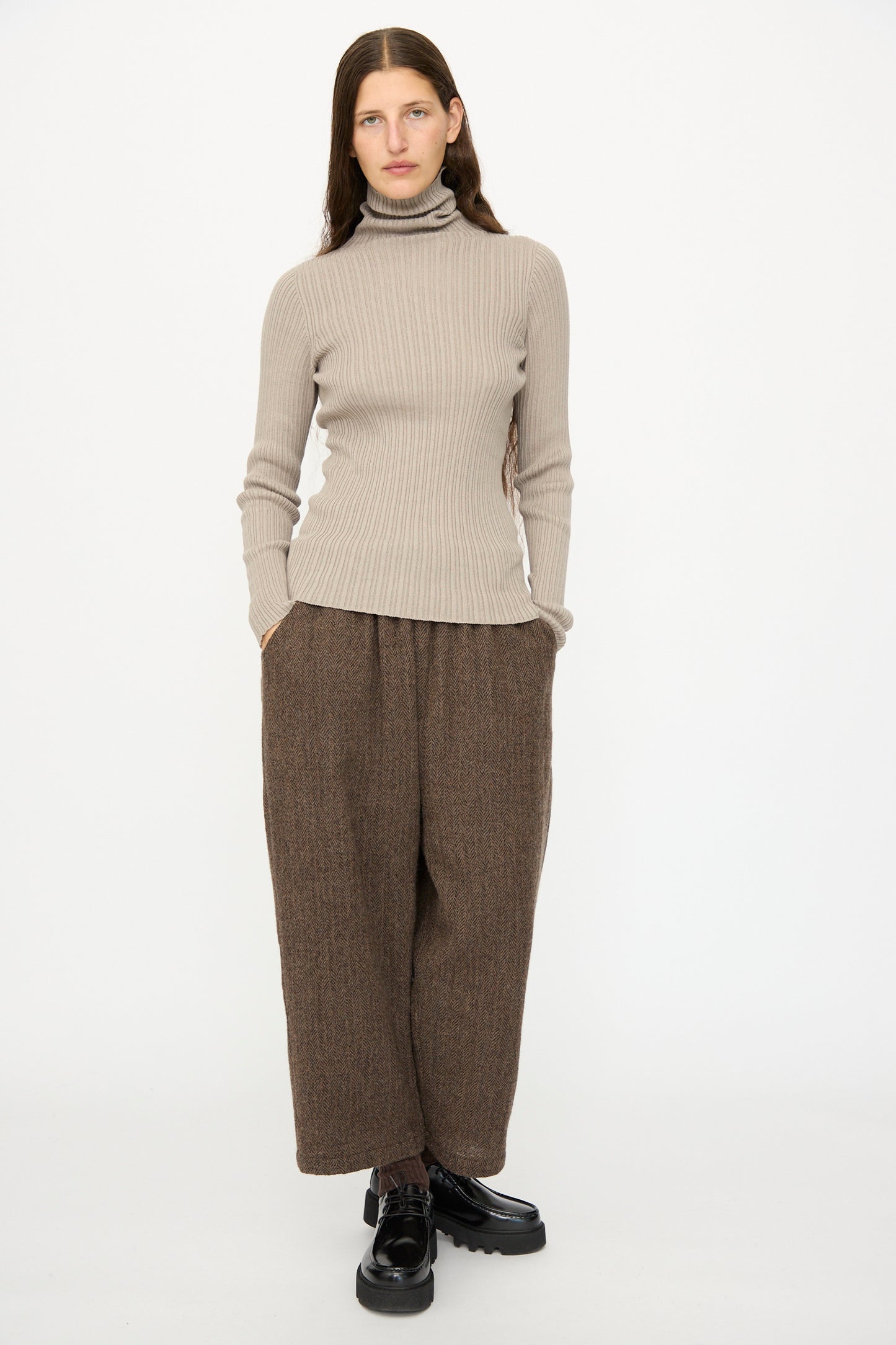 A person faces forward with hands in pockets, wearing the Ichi Antiquités Cotton Rib Knit Turtleneck in Mocha, loose brown pants, and black shoes against a plain white background.