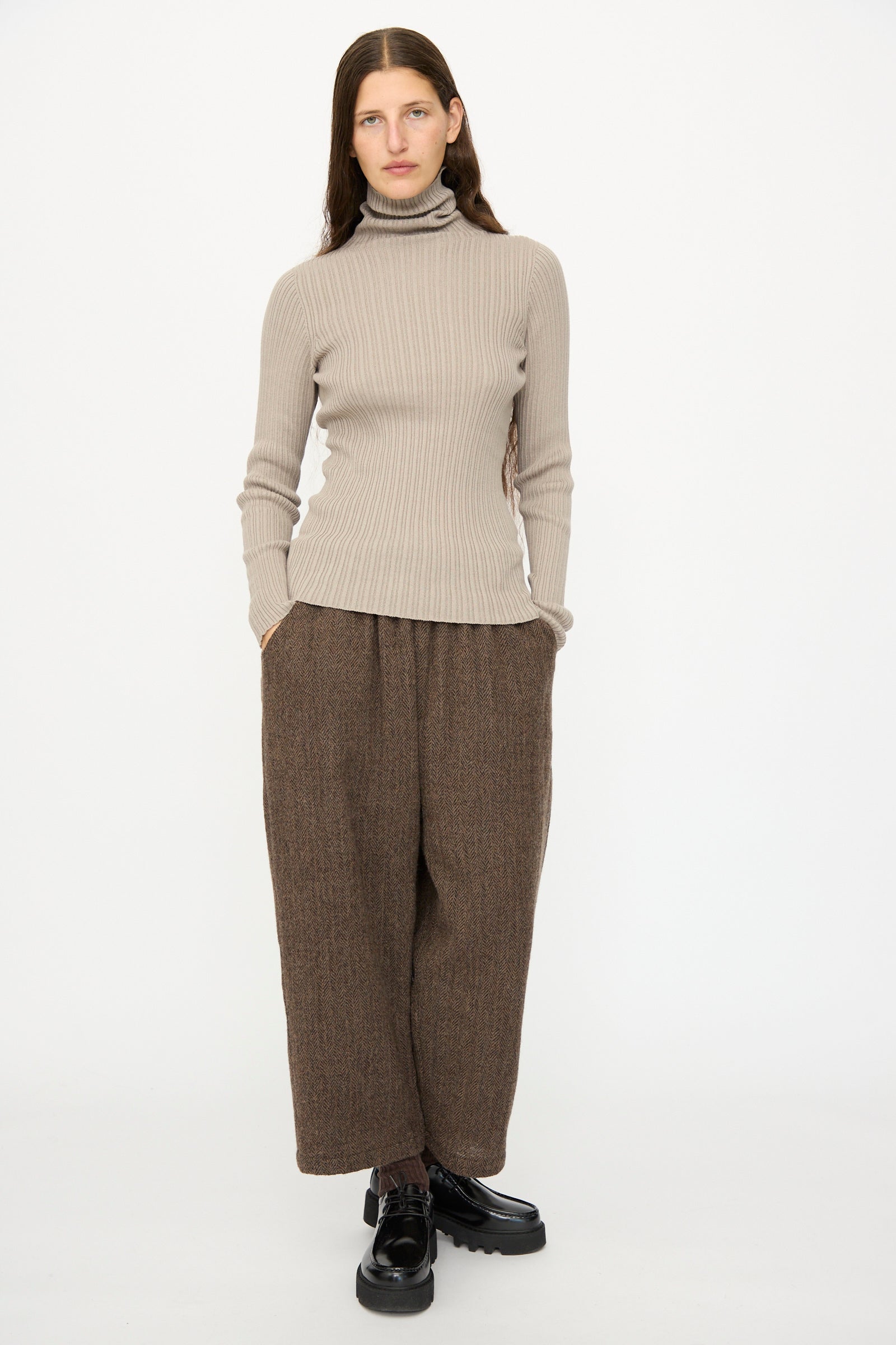 A person faces forward with hands in pockets, wearing the Ichi Antiquités Cotton Rib Knit Turtleneck in Mocha, loose brown pants, and black shoes against a plain white background.