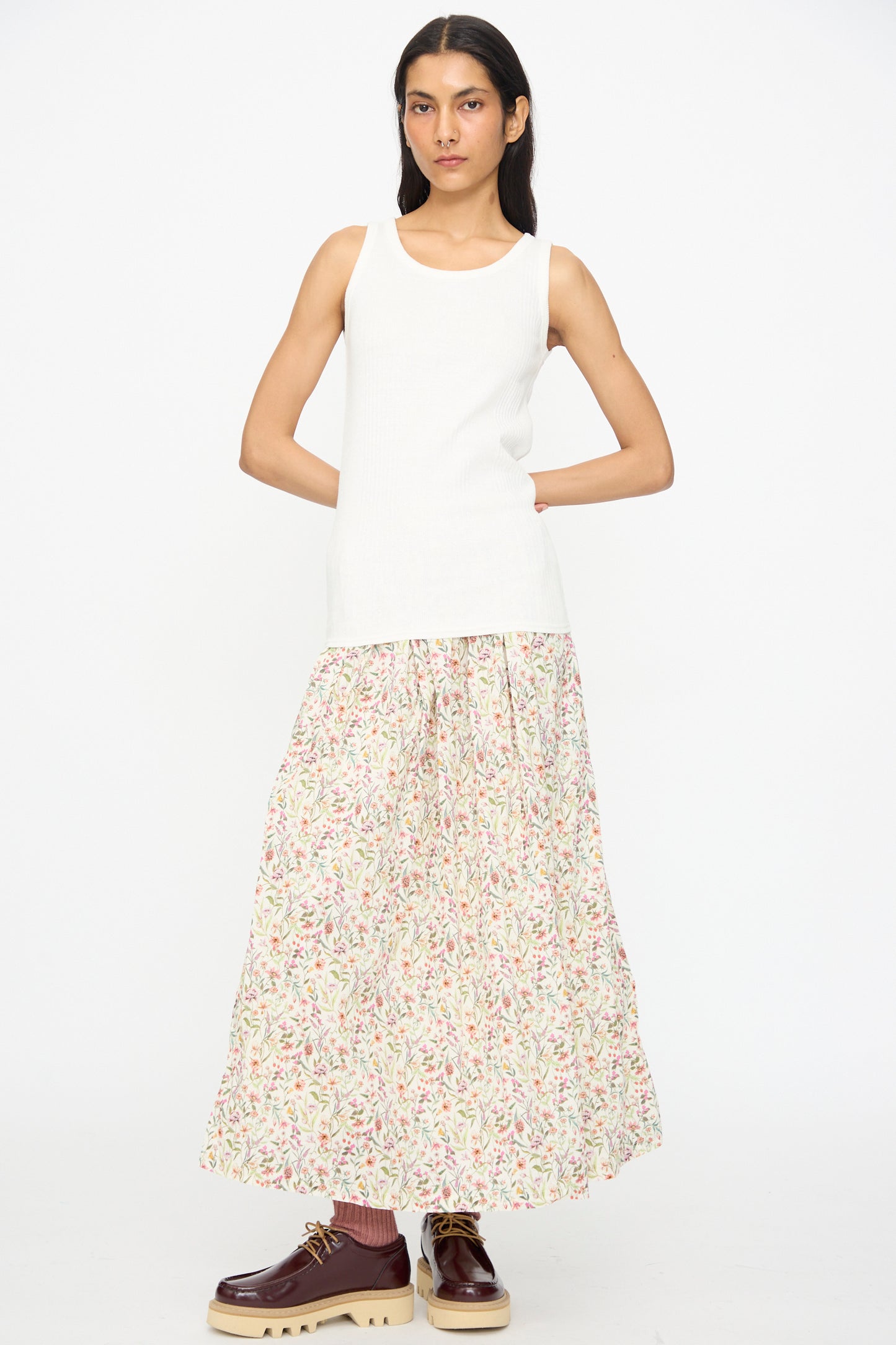 A person stands against a white background wearing the Ichi Antiquités Cotton Rib Tank Top in White, paired with a long floral skirt and brown lace-up shoes.