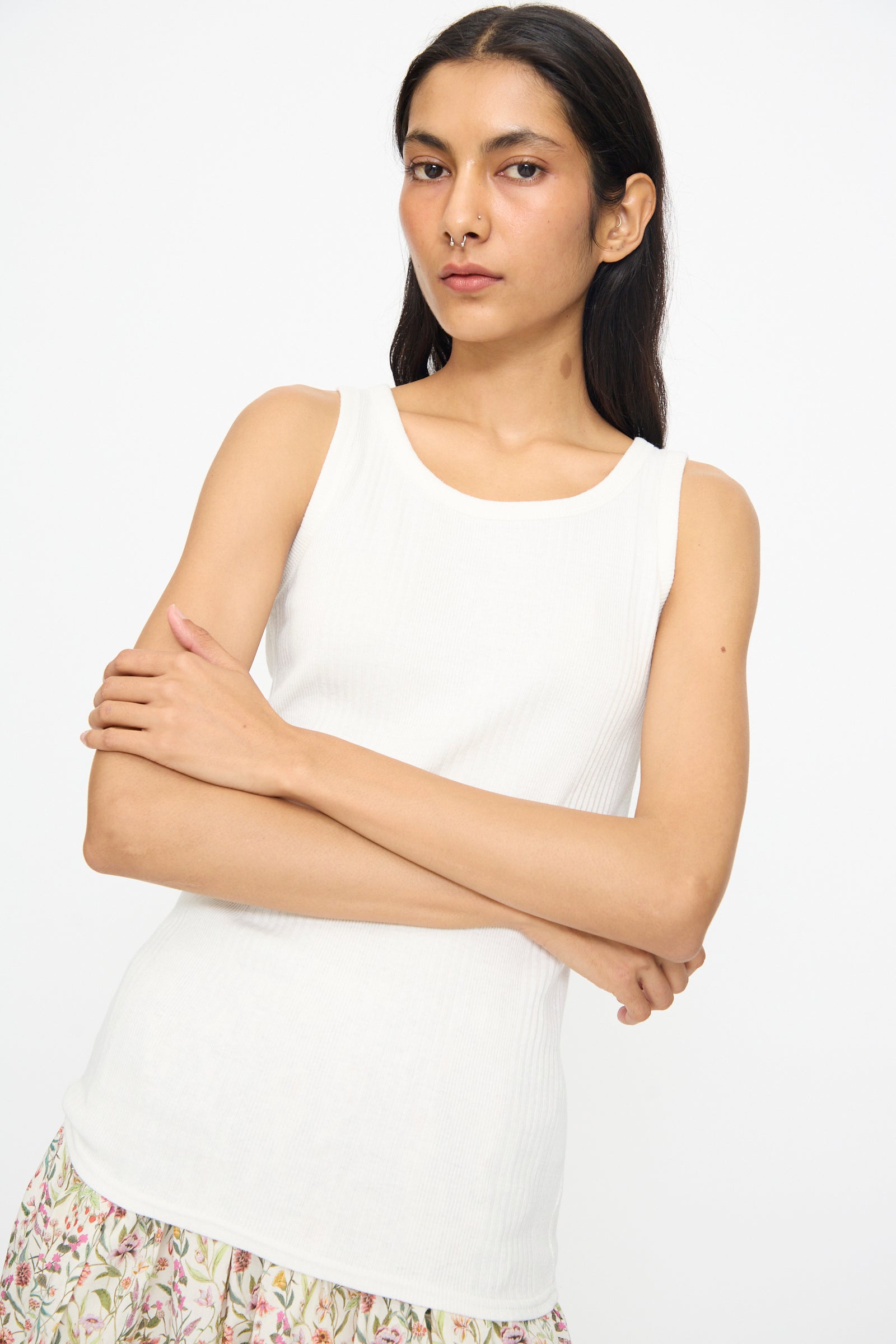 A person with long dark hair wears the Ichi Antiquités Cotton Rib Tank Top in White, paired with a patterned skirt, standing against a plain white background with arms crossed.