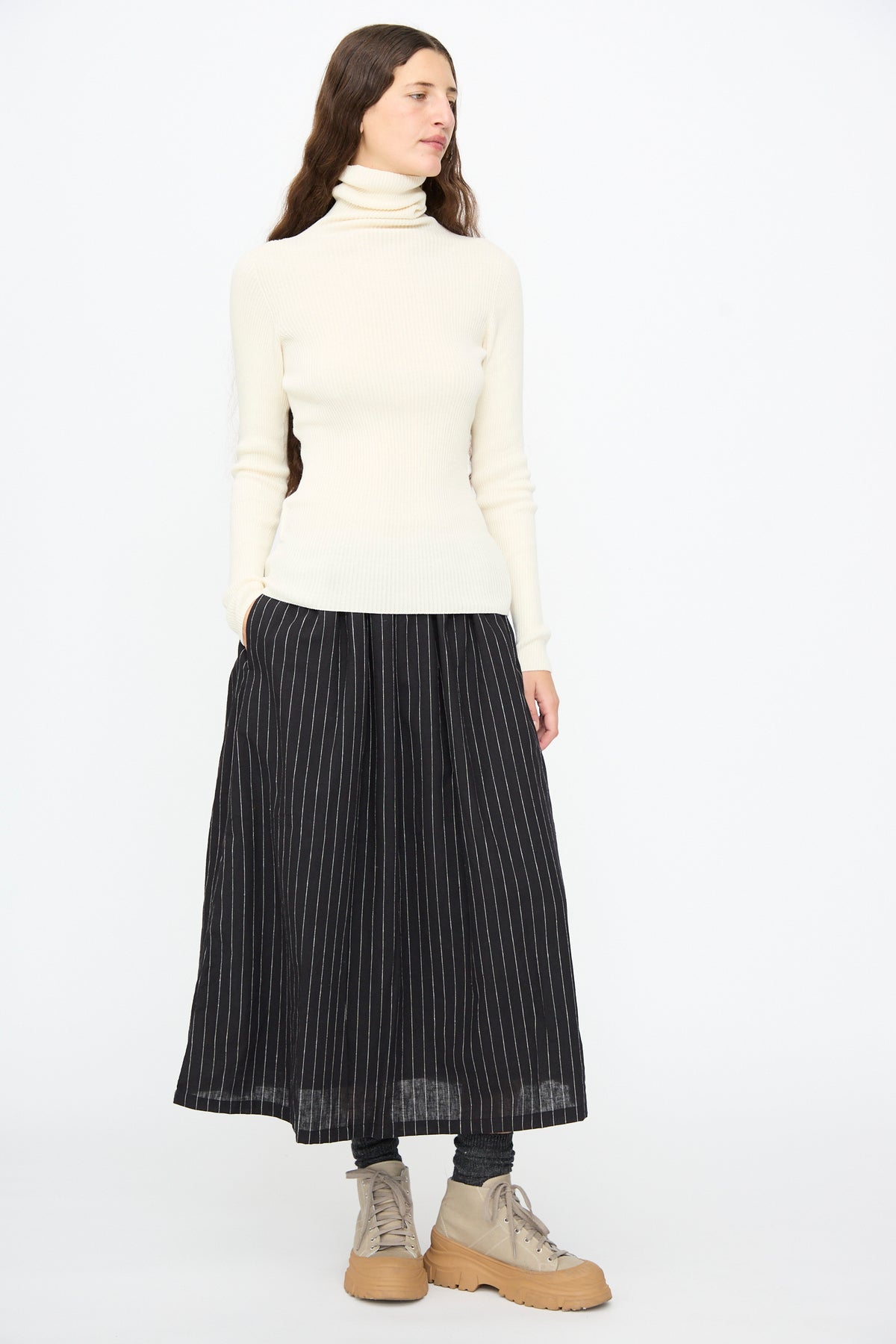 A person wearing an ivory Cotton Rib Turtleneck by Ichi Antiquités, a black pinstripe midi skirt, and tan platform sneakers.