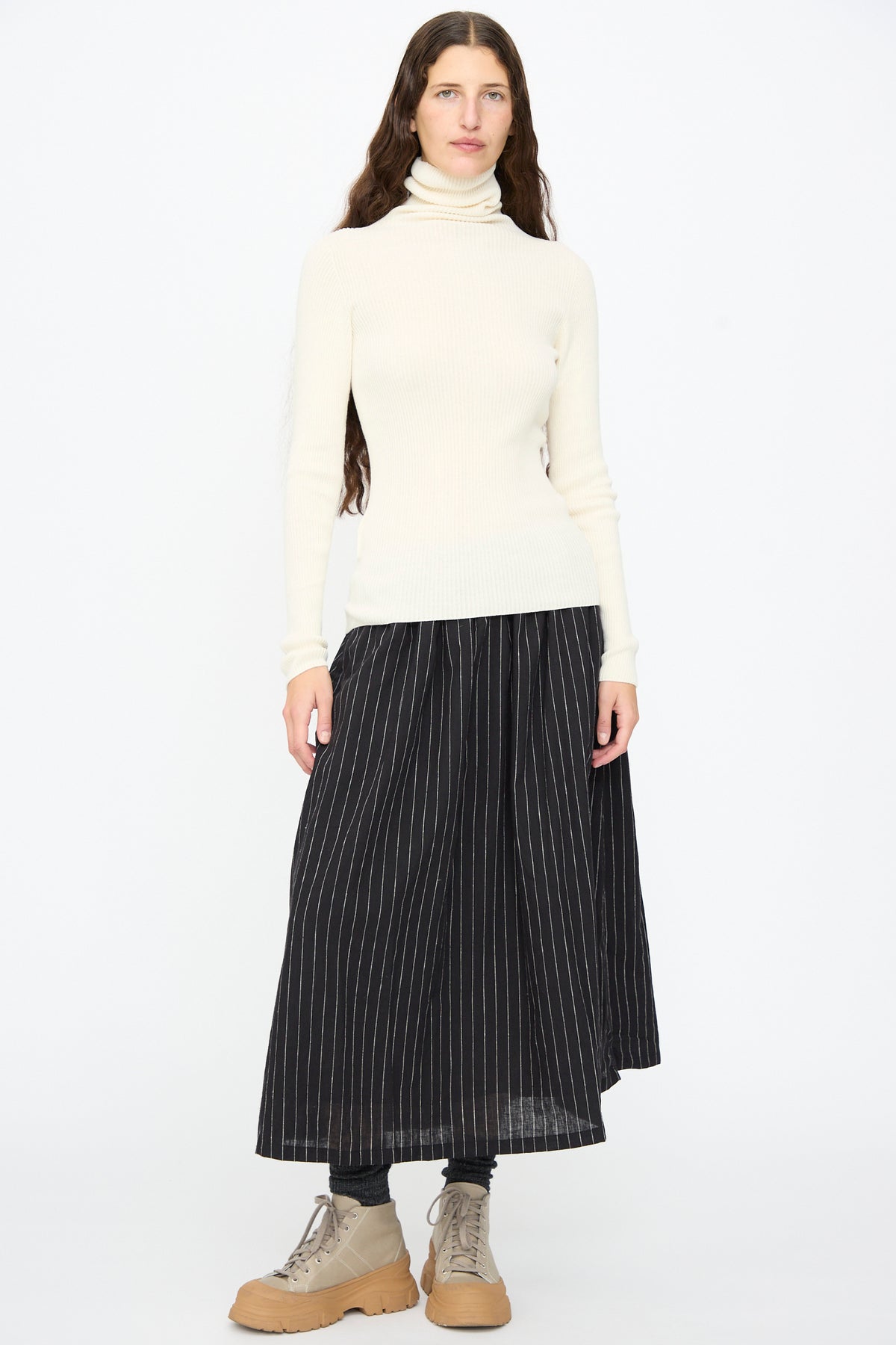 A person wearing an Ichi Antiquités Cotton Rib Turtleneck in Ivory, a black pinstriped midi skirt, and tan lace-up boots.