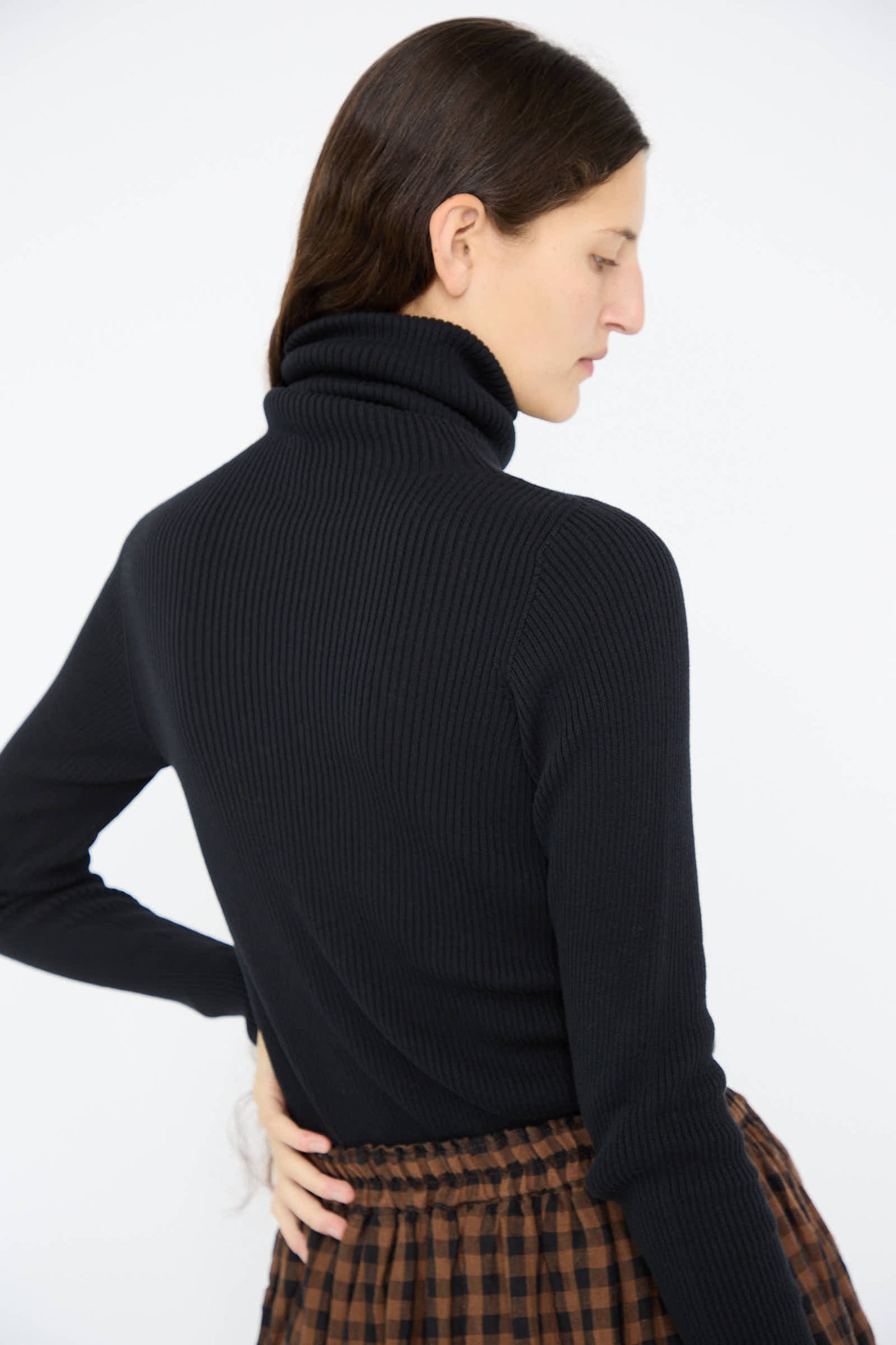 An individual wearing the Ichi Antiquités Cotton Rib Turtleneck in Black and a brown plaid skirt, seen from the side against a plain background.