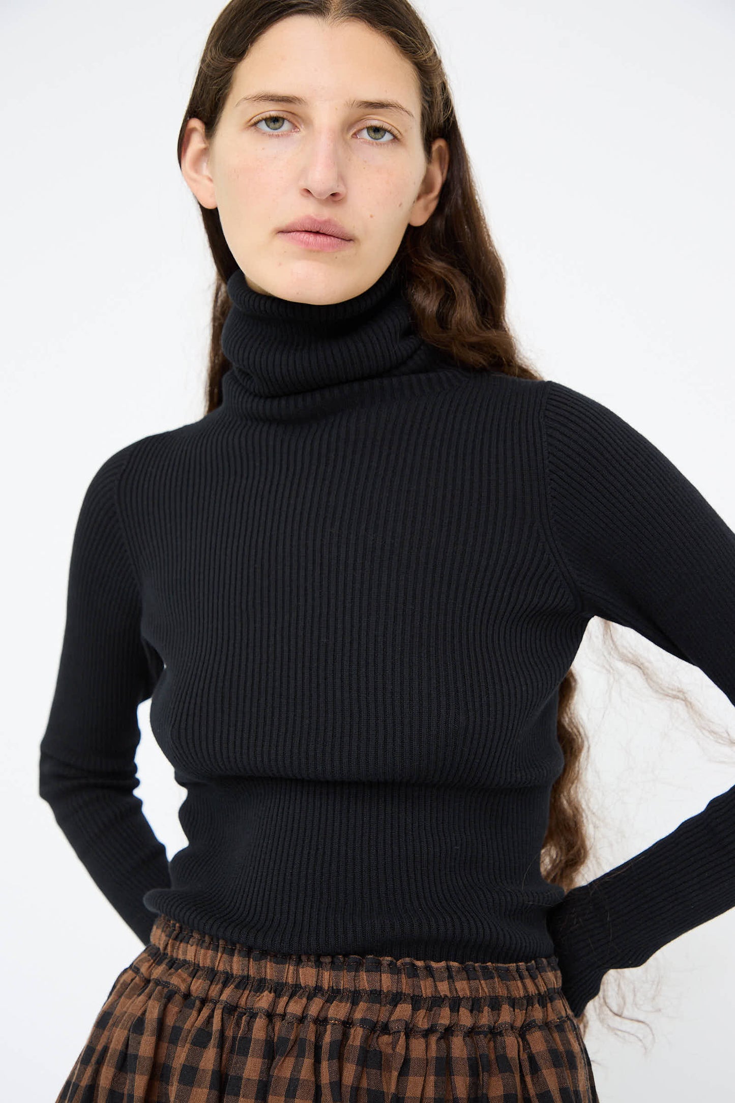 A person with long hair is wearing the Cotton Rib Turtleneck in Black by Ichi Antiquités and a plaid skirt against a plain background.