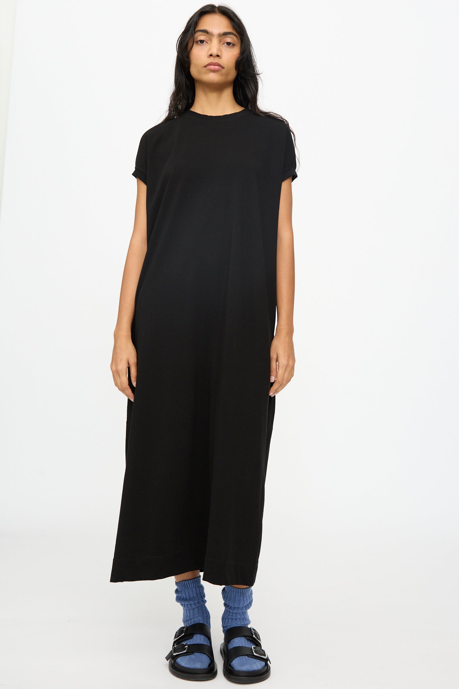 A person faces forward against a white background wearing the Ichi Antiquités Cotton Sleeveless Dress in Black, styled with blue socks and black sandals.