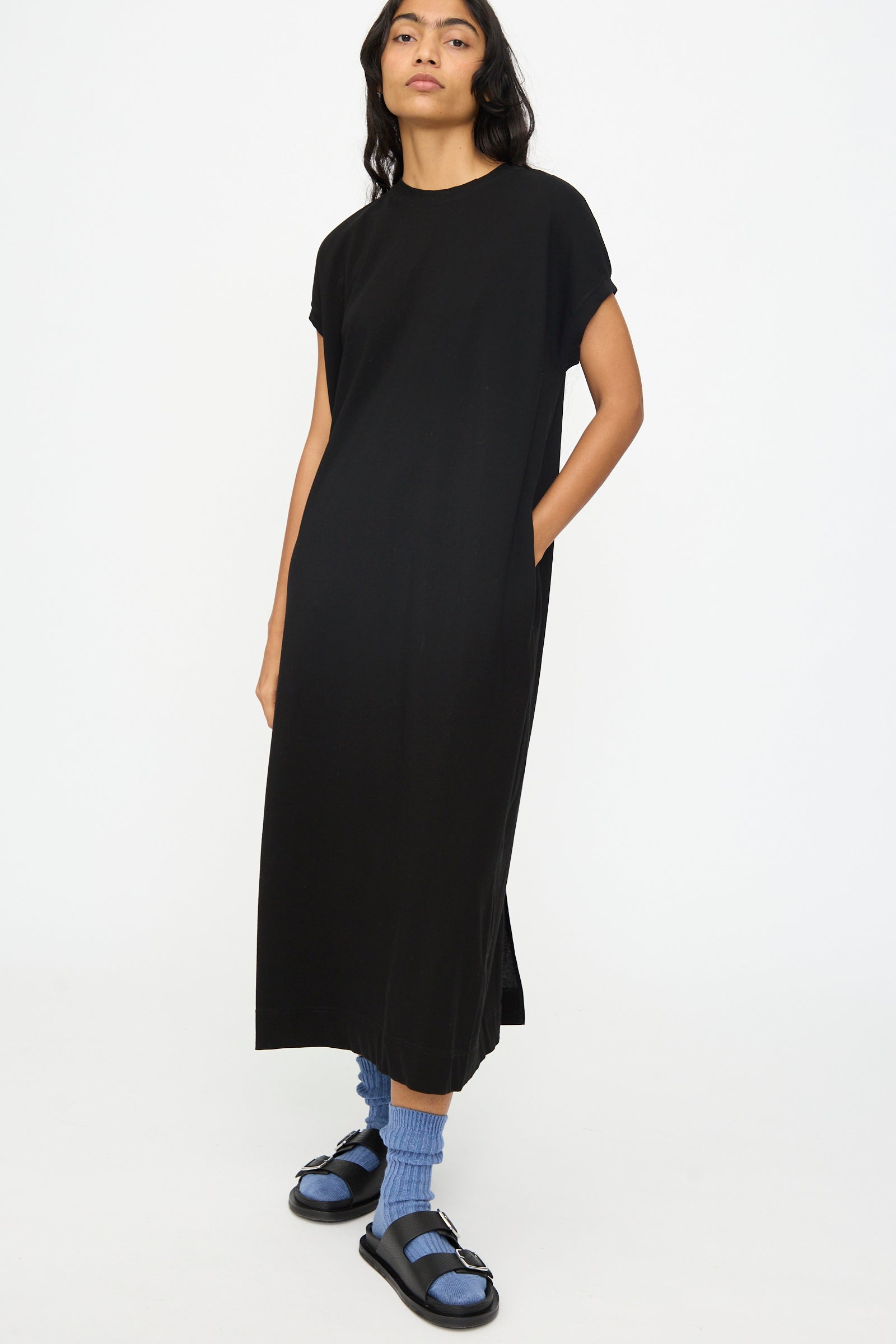A person stands against a plain white background wearing the Ichi Antiquités Cotton Sleeveless Dress in Black, paired with blue socks and black sandals.