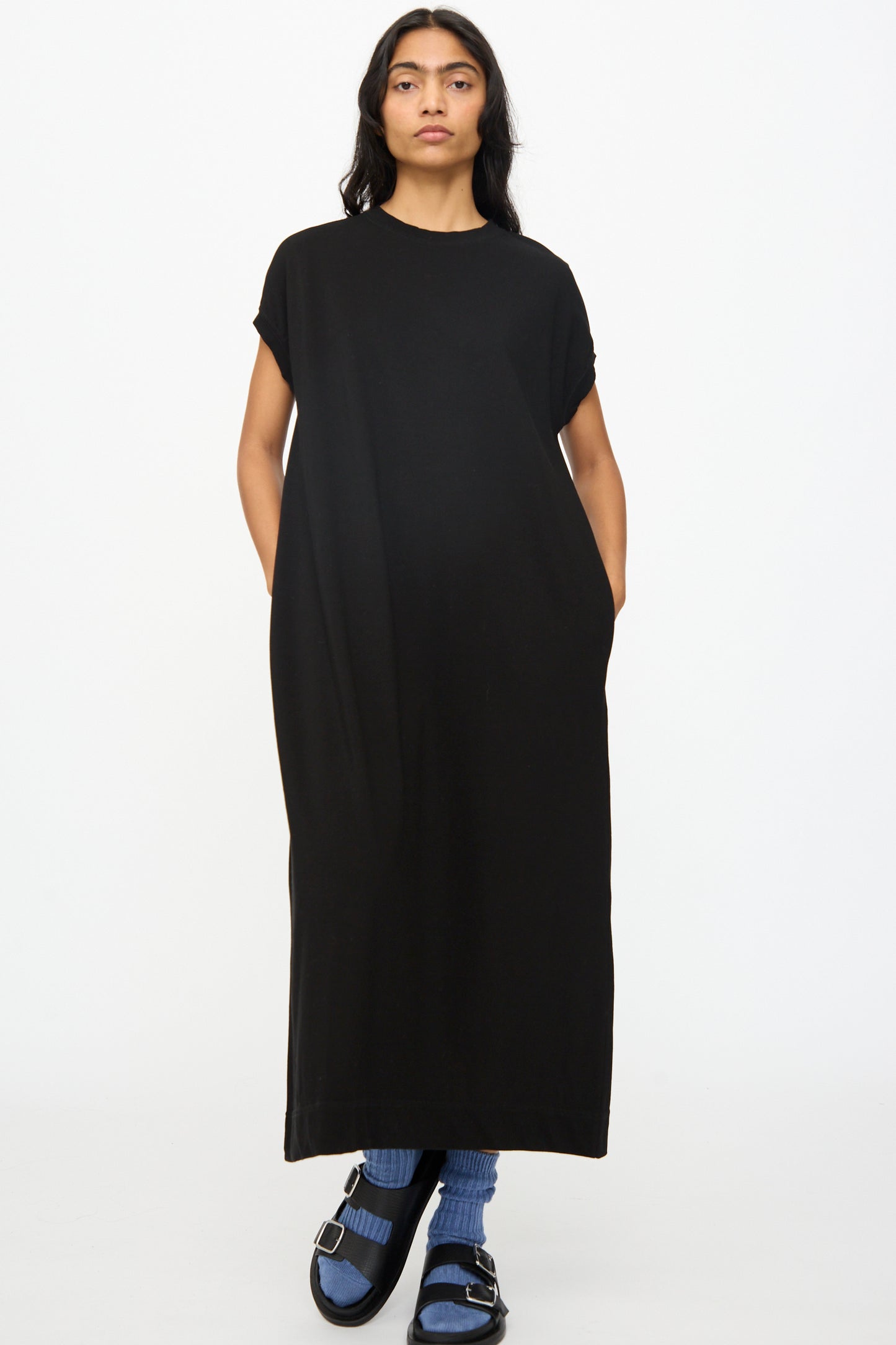 A person faces forward wearing the Ichi Antiquités Cotton Sleeveless Dress in Black, paired with blue socks and black sandals, against a plain white background.