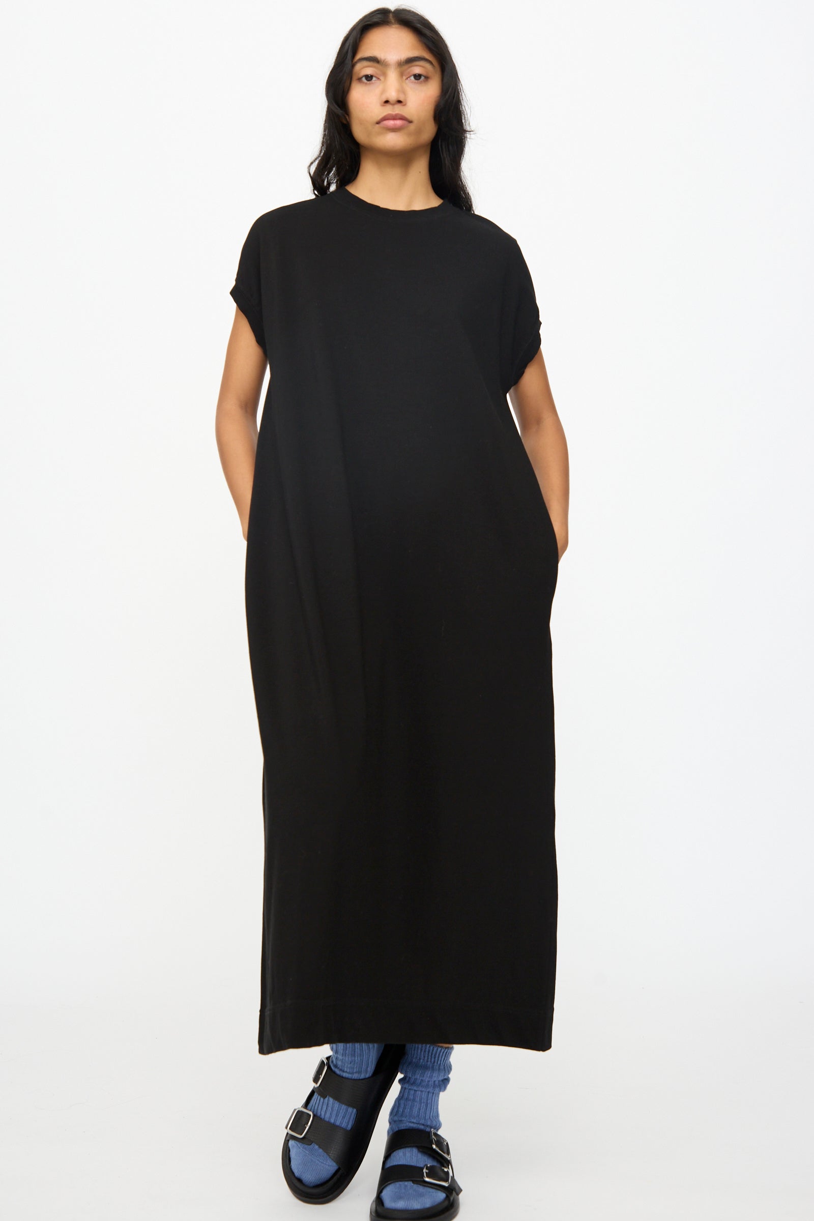 A person faces forward wearing the Ichi Antiquités Cotton Sleeveless Dress in Black, paired with blue socks and black sandals, against a plain white background.