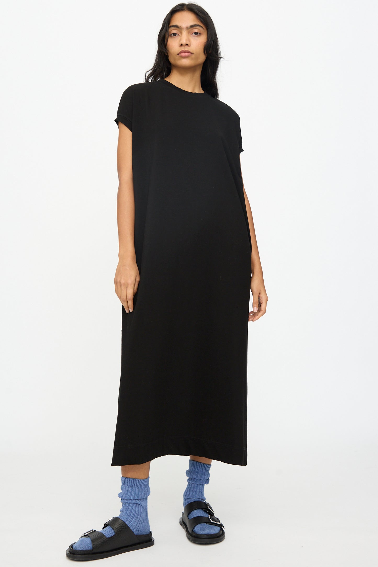 A person stands against a plain background wearing the Ichi Antiquités Cotton Sleeveless Dress in Black, styled as a pullover, paired with blue socks and black sandals.