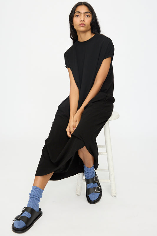 A person with long dark hair sits on a white stool, wearing the Cotton Sleeveless Dress in Black by Ichi Antiquités, paired with blue socks and black sandals against a plain white background.