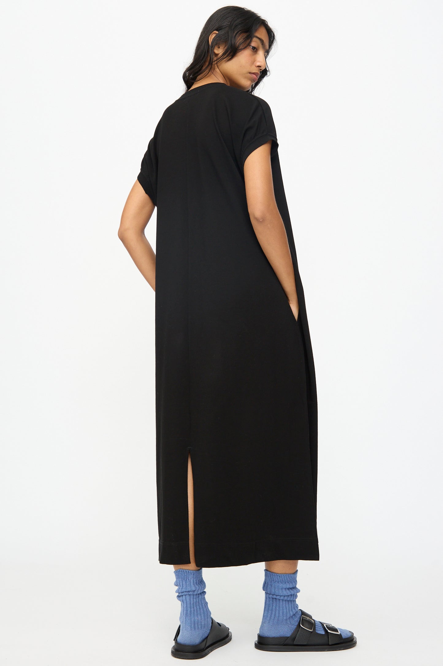 A woman faces away from the camera, wearing the Ichi Antiquités Cotton Sleeveless Dress in Black, paired with blue socks and black sandals, set against a plain white background.