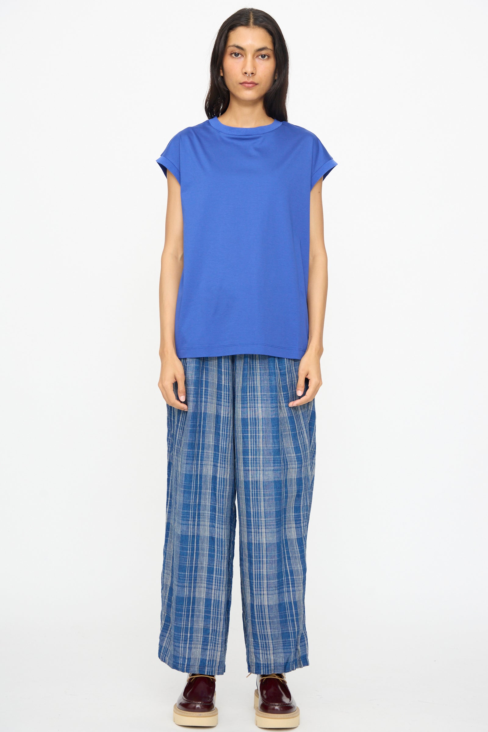 A person stands facing forward against a plain white background, wearing the Ichi Antiquités Cotton Sleeveless Pullover in Blue, blue plaid wide-leg pants, and burgundy shoes.