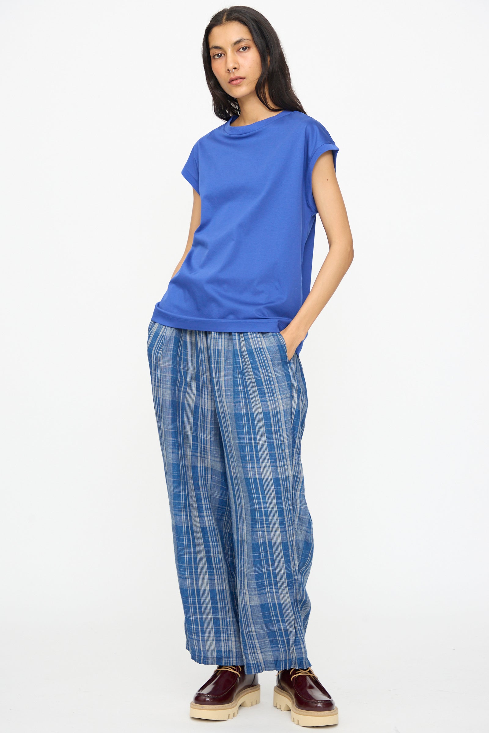 Person standing against a white background, wearing the Ichi Antiquités Cotton Sleeveless Pullover in Blue, blue plaid wide-leg pants, and burgundy platform shoes with cream soles. Hands are in their pockets.