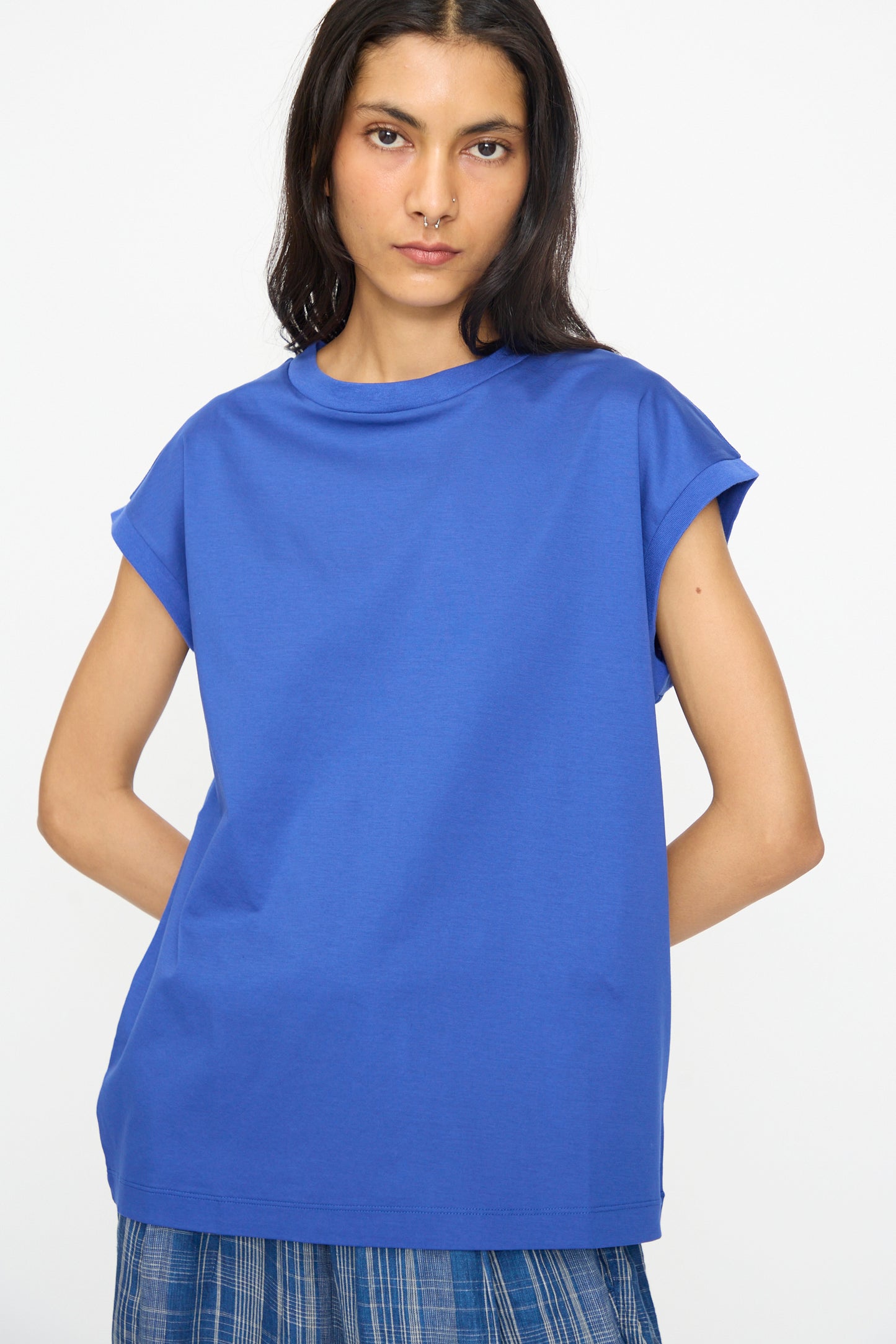 A person with long dark hair wears the Ichi Antiquités Cotton Sleeveless Pullover in Blue, paired with a blue plaid skirt, standing against a plain white background.