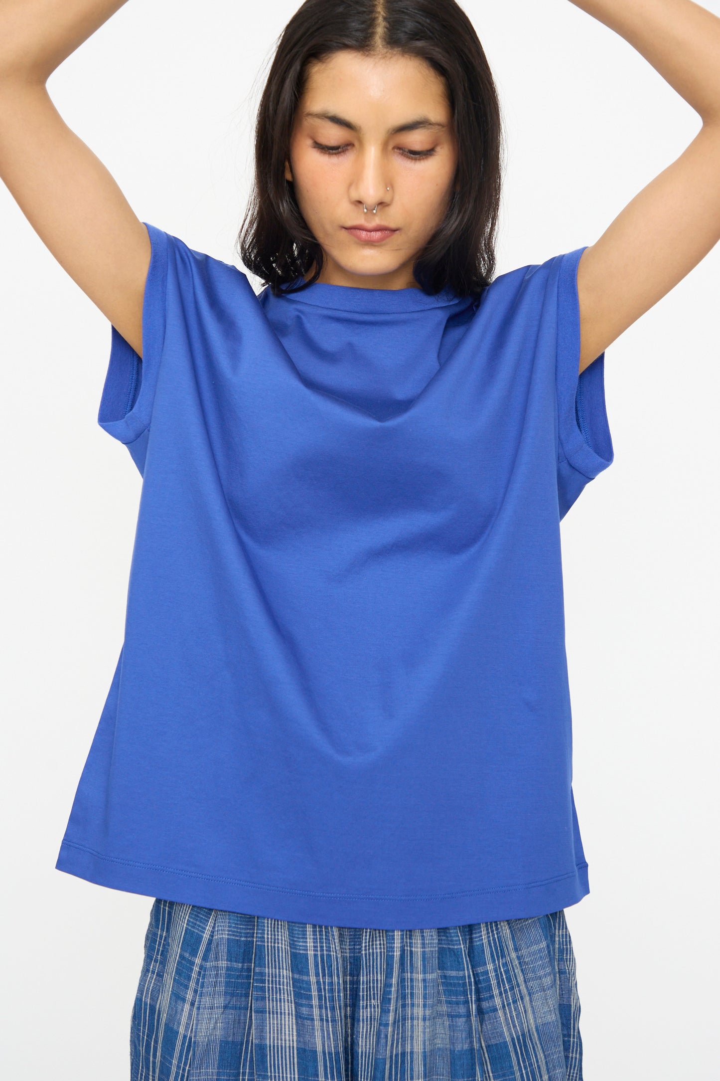 A person wears the Ichi Antiquités Cotton Sleeveless Pullover in Blue with a plaid skirt, standing with arms raised against a plain white background.