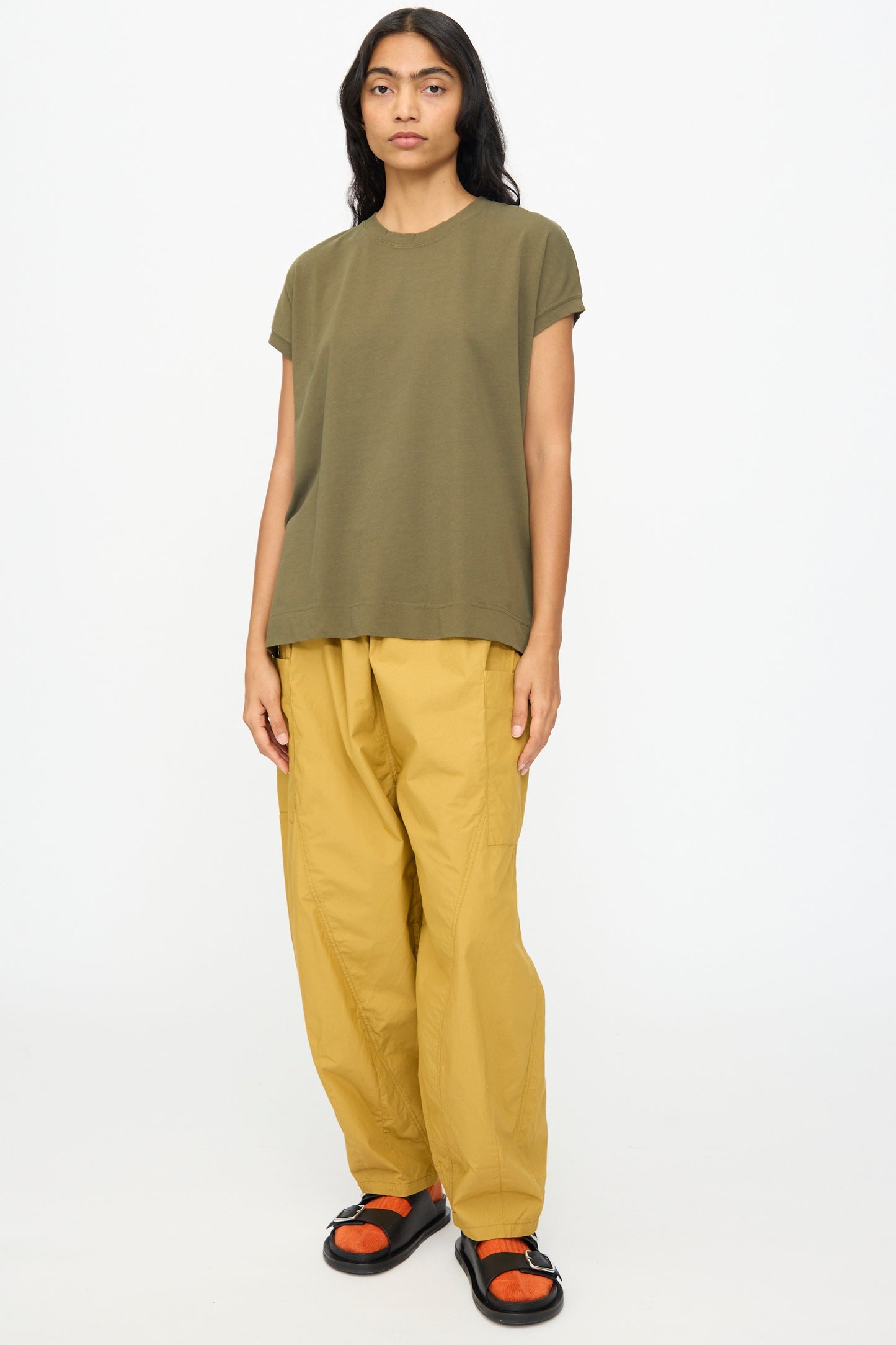 A person faces forward wearing the Ichi Antiquités Cotton Sleeveless Tee in Khaki, paired with loose yellow pants and black sandals with orange accents, set against a plain white background.
