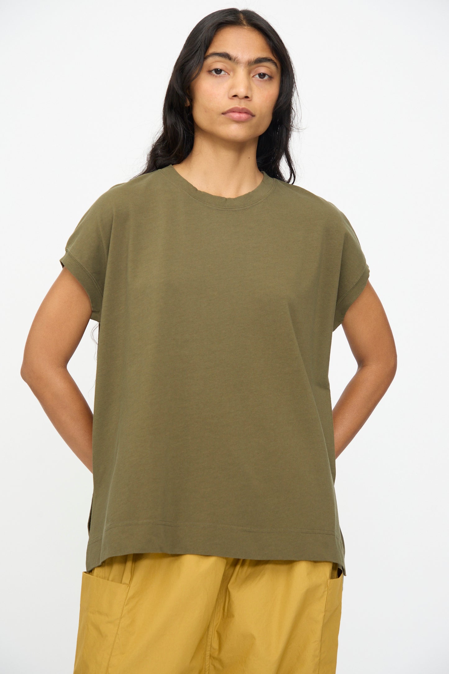 A person with long dark hair wears the Ichi Antiquités Cotton Sleeveless Tee in Khaki, paired with mustard yellow pants, and stands against a plain white background.