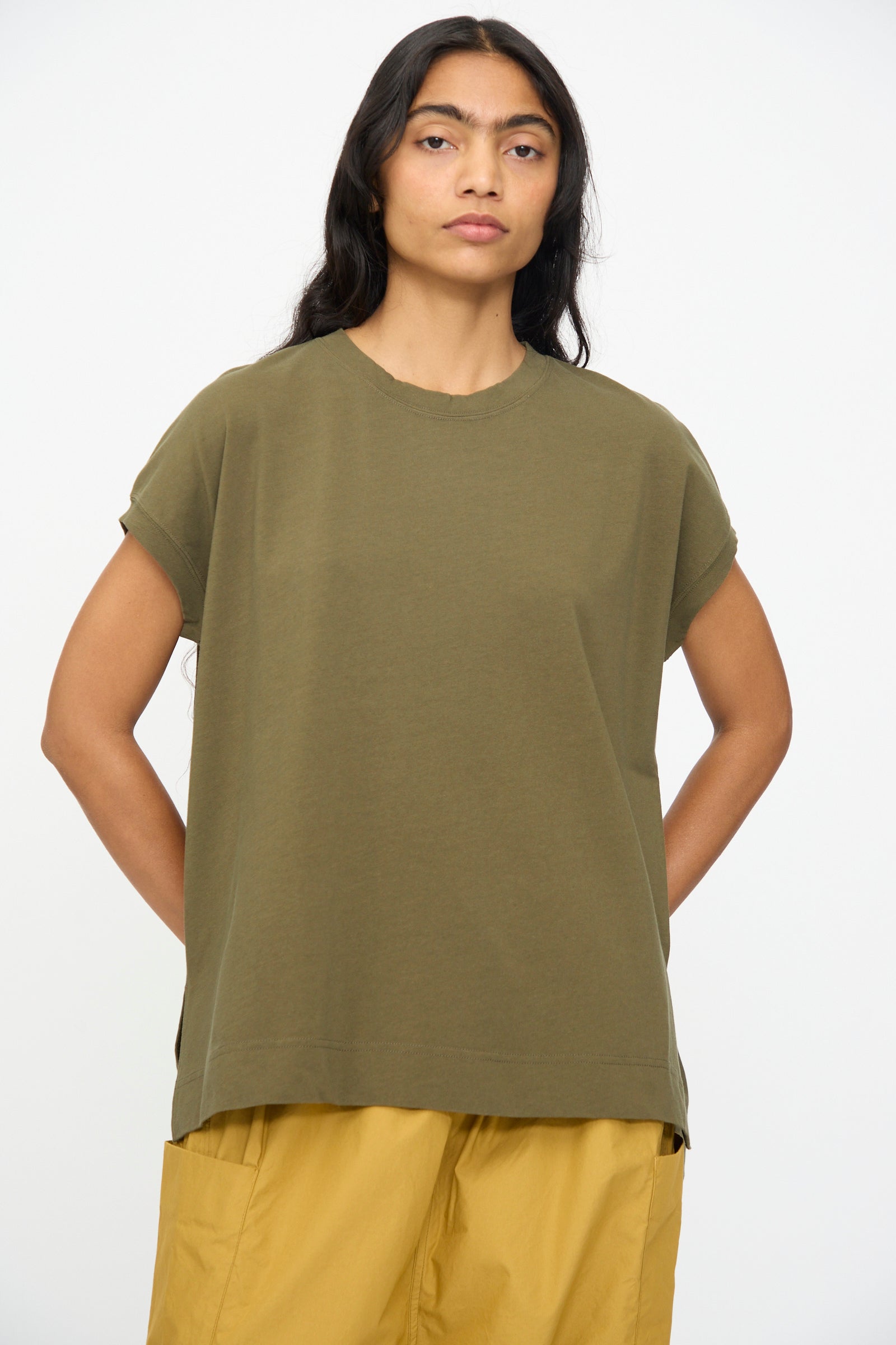 A person with long dark hair wears the Ichi Antiquités Cotton Sleeveless Tee in Khaki, paired with mustard yellow pants, and stands against a plain white background.