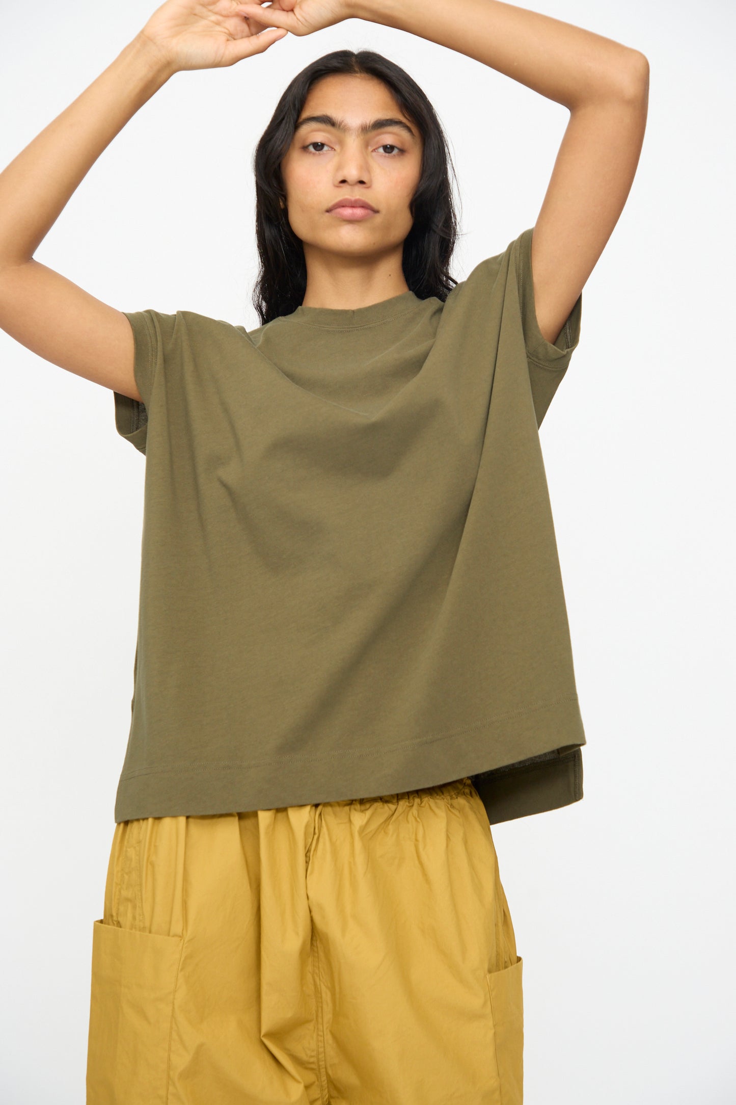 A woman stands with arms raised, wearing the Ichi Antiquités Cotton Sleeveless Tee in Khaki and mustard yellow pants with large pockets, set against a plain light background.