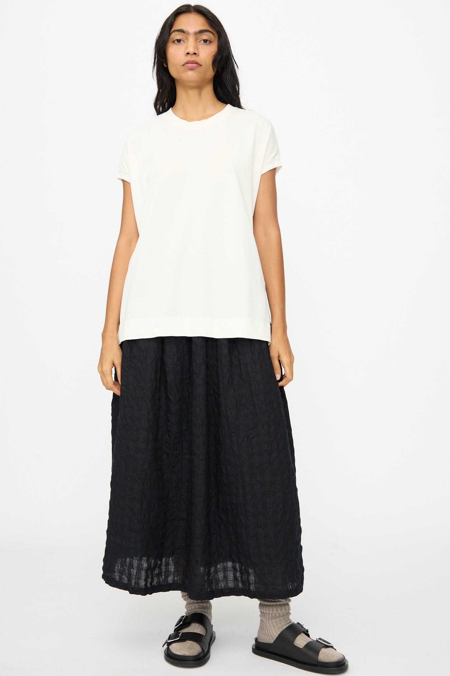 A person stands facing forward wearing the Ichi Antiquités Cotton Sleeveless Tee in White, a long black textured skirt, gray socks, and black sandals against a plain white background.