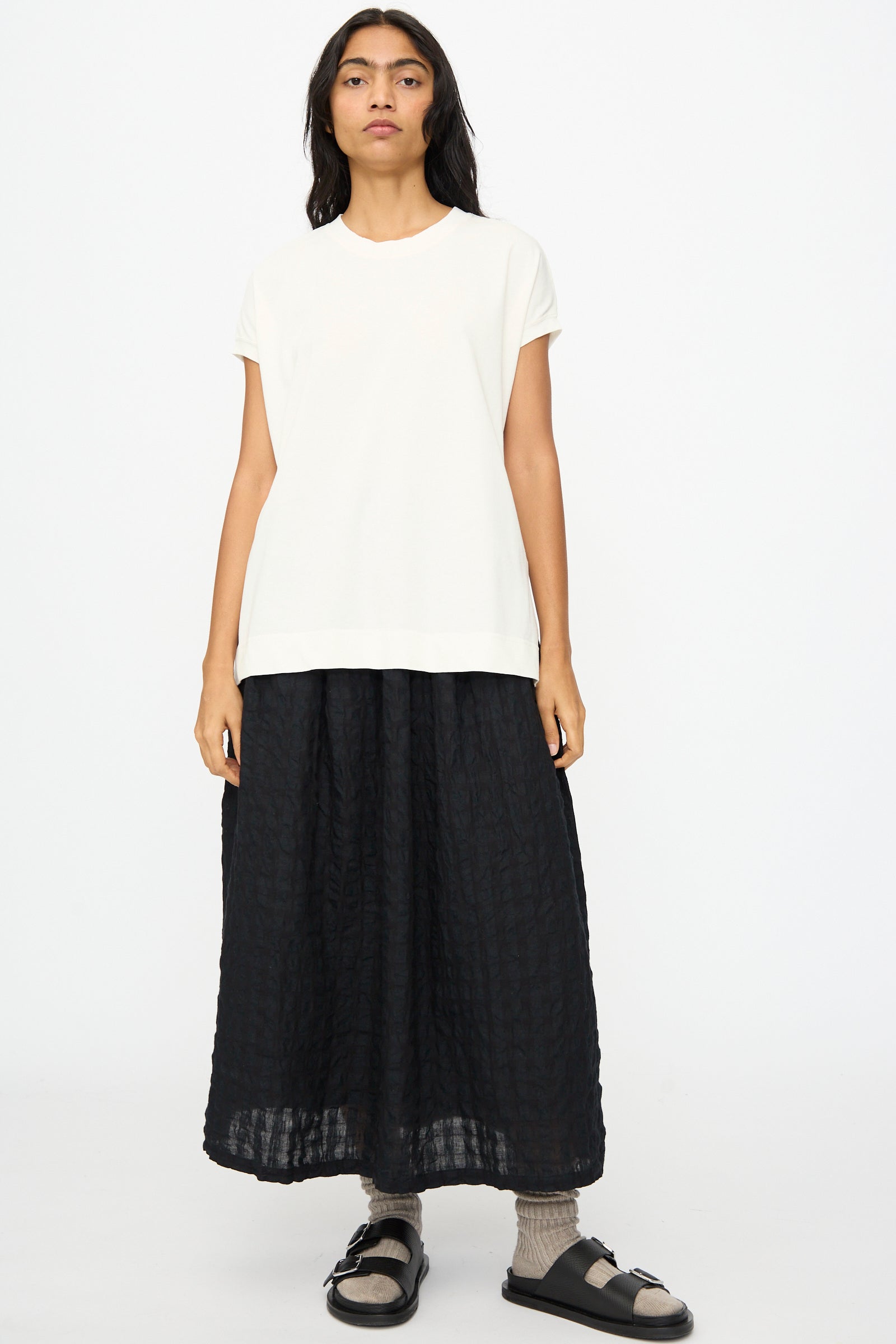 A person stands facing forward wearing the Ichi Antiquités Cotton Sleeveless Tee in White, a long black textured skirt, gray socks, and black sandals against a plain white background.