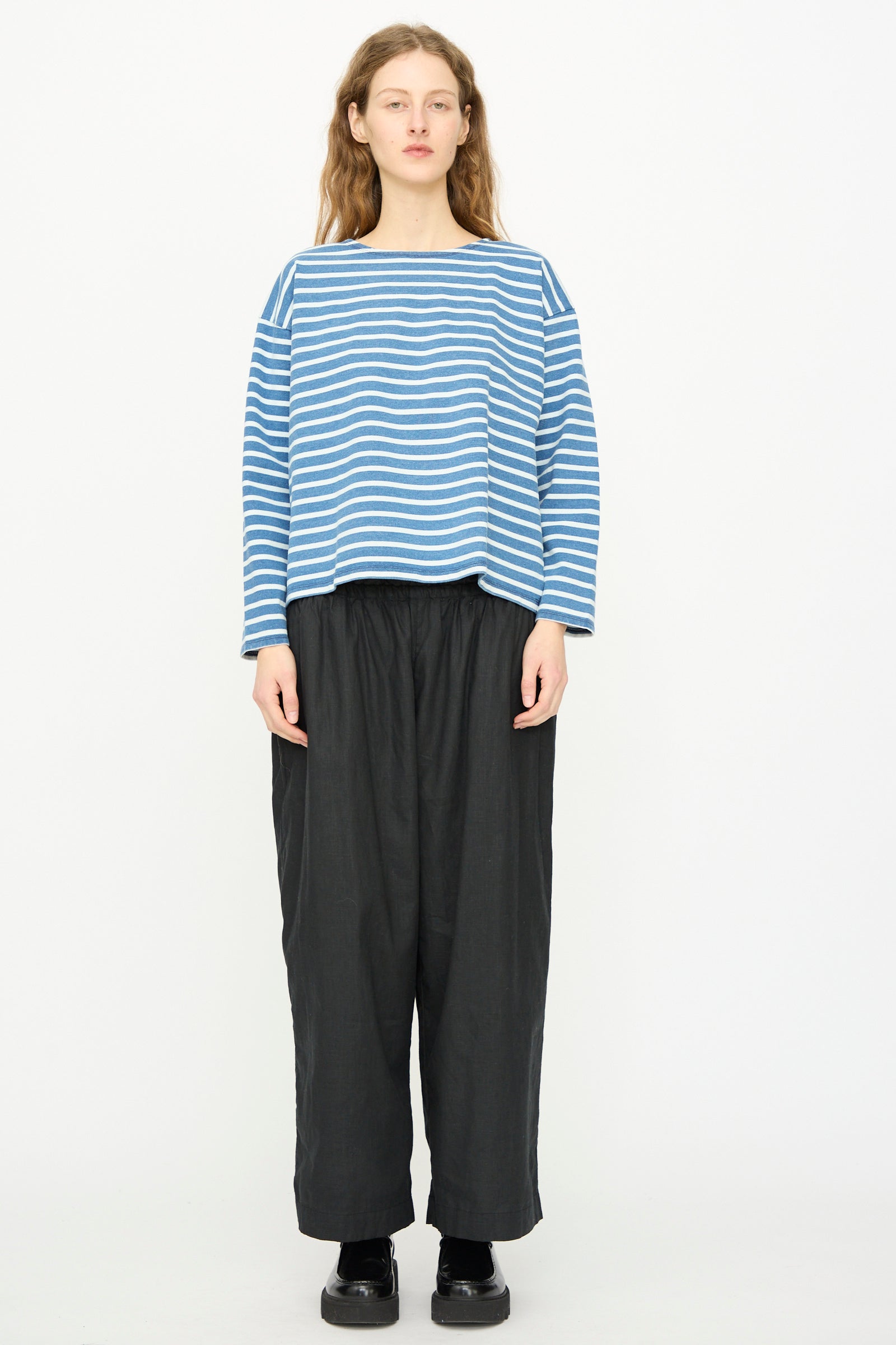 A person stands against a plain background wearing the Ichi Antiquités Cotton Stripe Pullover in Blue and White, paired with loose black pants and black shoes.