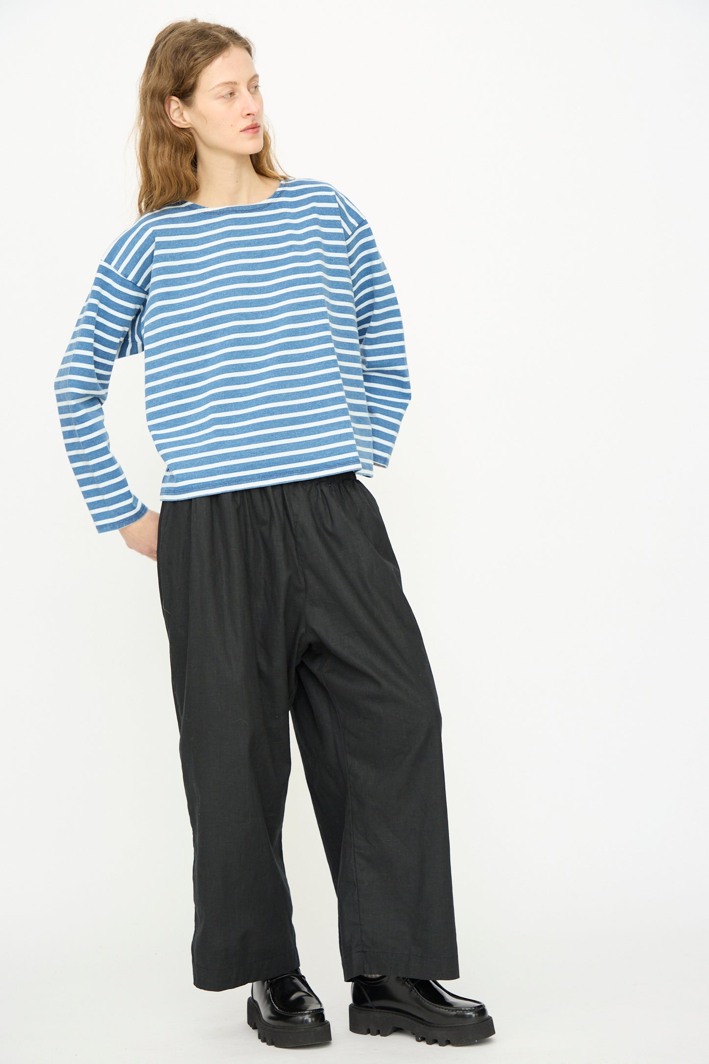 Against a plain white background, a person wears the Ichi Antiquités Cotton Stripe Pullover in Blue and White, paired with loose black pants and black platform shoes.