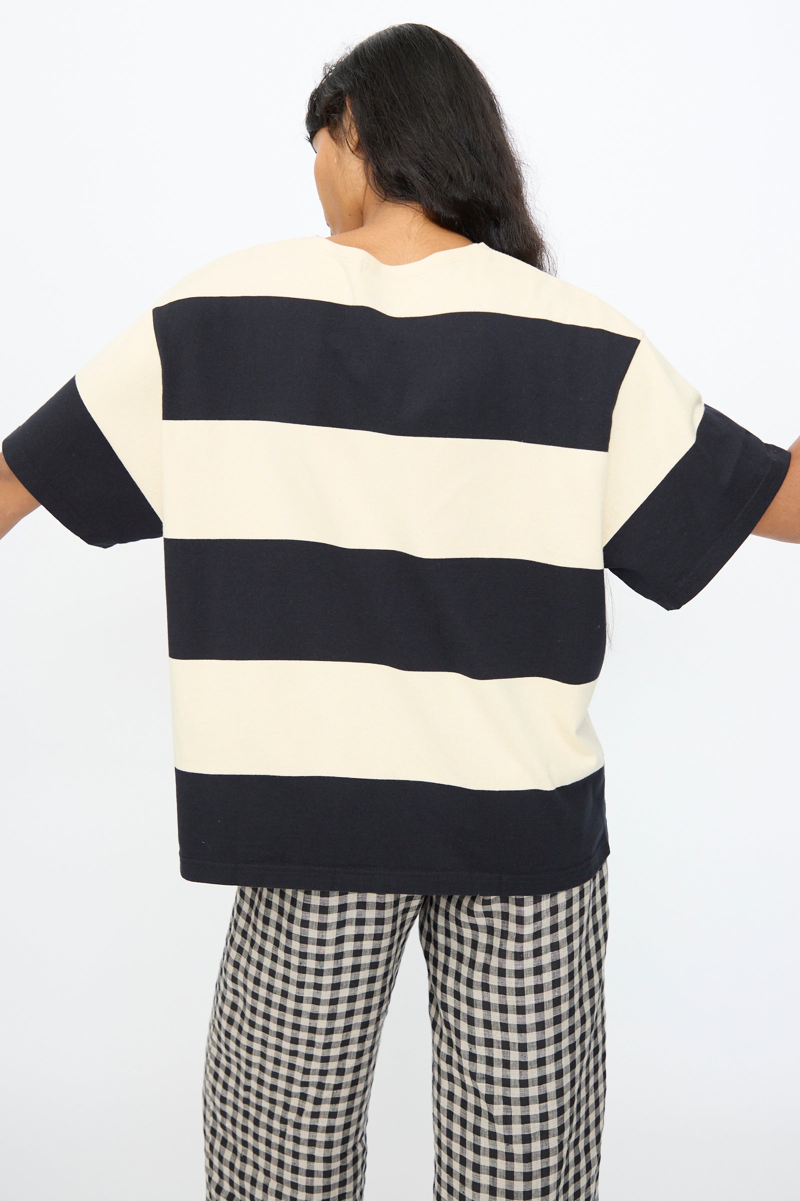 A person faces away with arms slightly extended, wearing the Ichi Antiquités Cotton Stripe Pullover in Natural and Black—a loose-fit Japanese-made cotton top styled with black-and-white checkered pants.