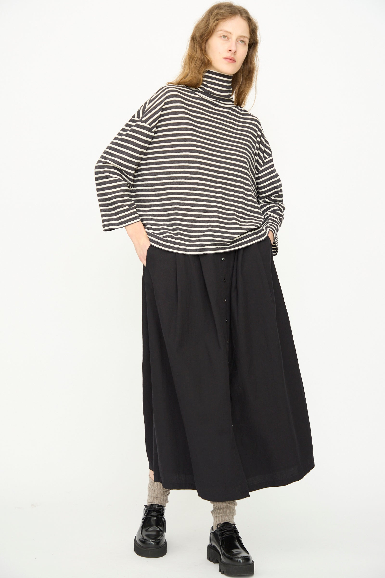 Model wears the Ichi Antiquités Cotton Stripe Turtleneck in Charcoal with a long black skirt, beige socks, and black platform shoes, standing hands in pockets against a white background.