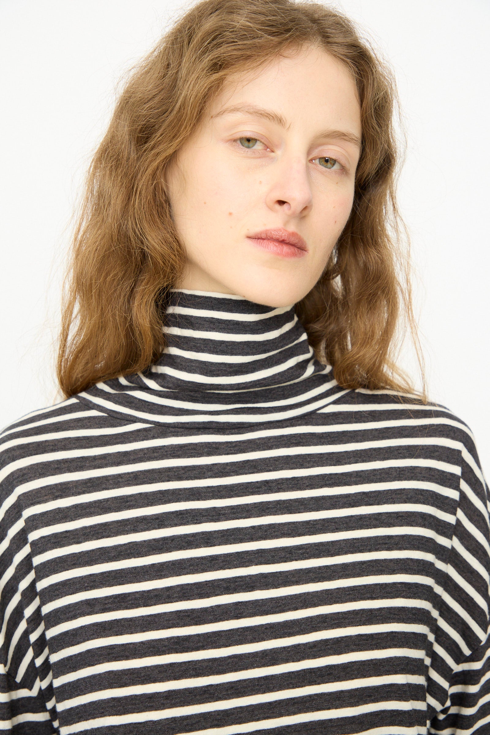A person with long, wavy hair wears the Ichi Antiquités Cotton Stripe Turtleneck in Charcoal, standing against a plain light background.