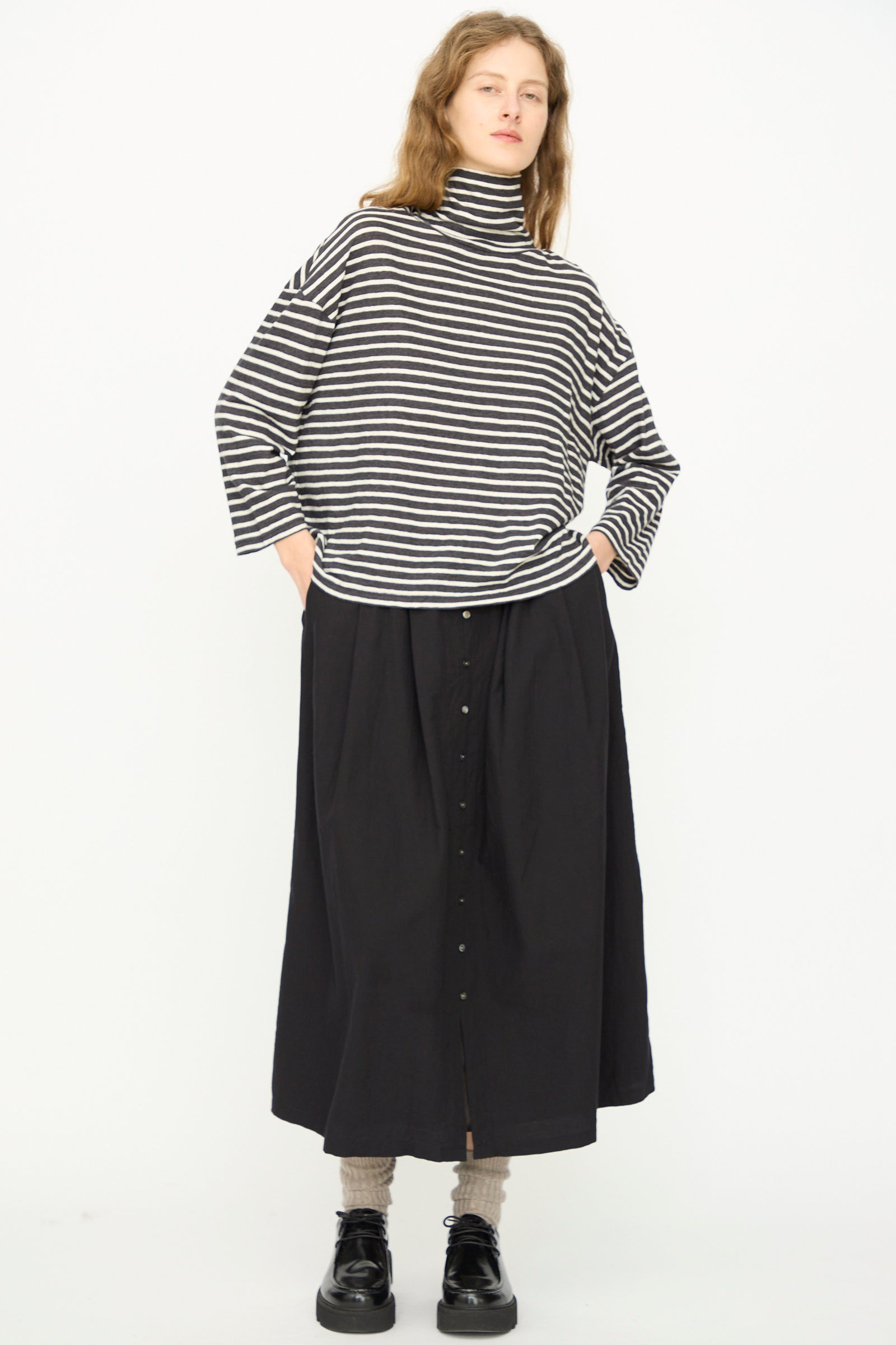 A person stands against a white background wearing the Ichi Antiquités Cotton Stripe Turtleneck in Charcoal, paired with a long black buttoned skirt, gray socks, and black shoes. Hands are in pockets. 
