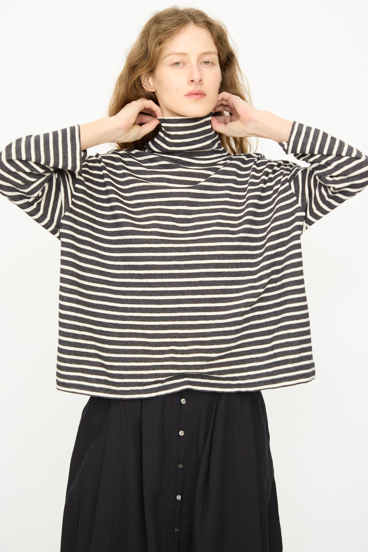 A person is wearing the Ichi Antiquités Cotton Stripe Turtleneck in Charcoal with a black buttoned skirt, standing against a plain white background.