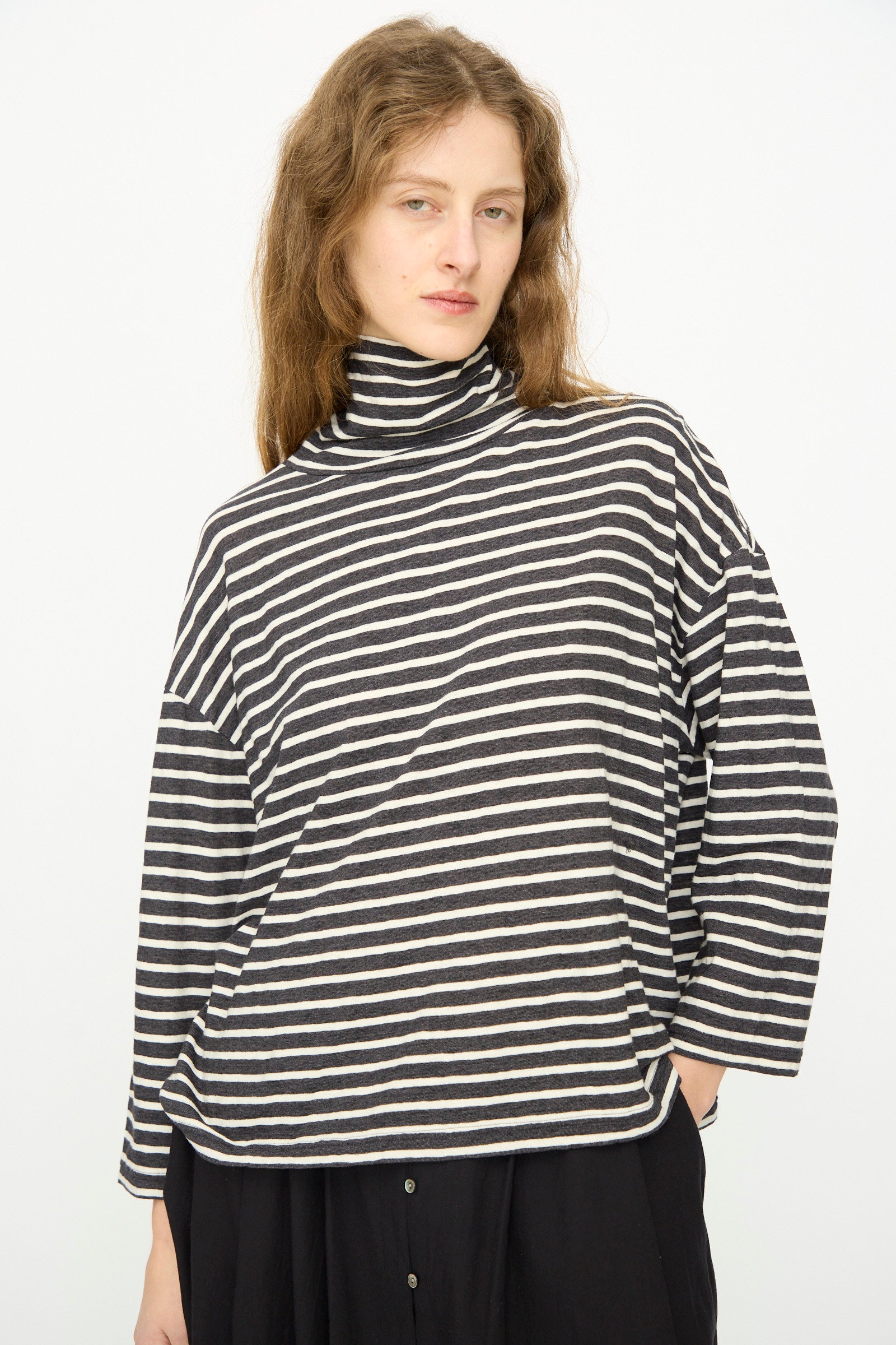 Model wearing the Ichi Antiquités Cotton Stripe Turtleneck in Charcoal, paired with a black buttoned skirt, stands against a plain white background.