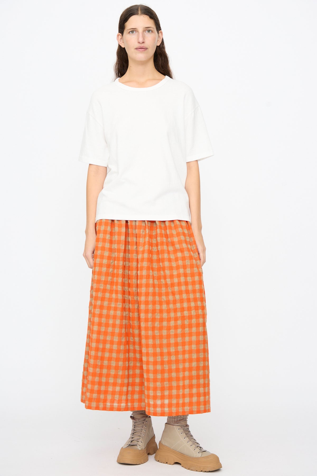 A person wears an oversized, made in Japan white Cotton T-Shirt in White, an orange and beige checkered midi skirt, and tan lace-up boots.