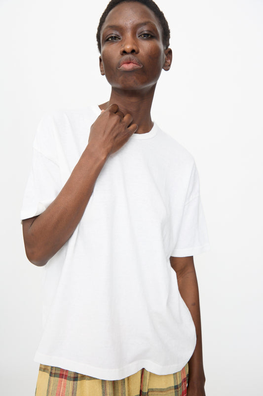 Person stands against a plain background, wearing the Ichi Antiquités Cotton T-Shirt in White—an oversized Japanese-made top—paired with yellow plaid pants and lightly touching their neck.