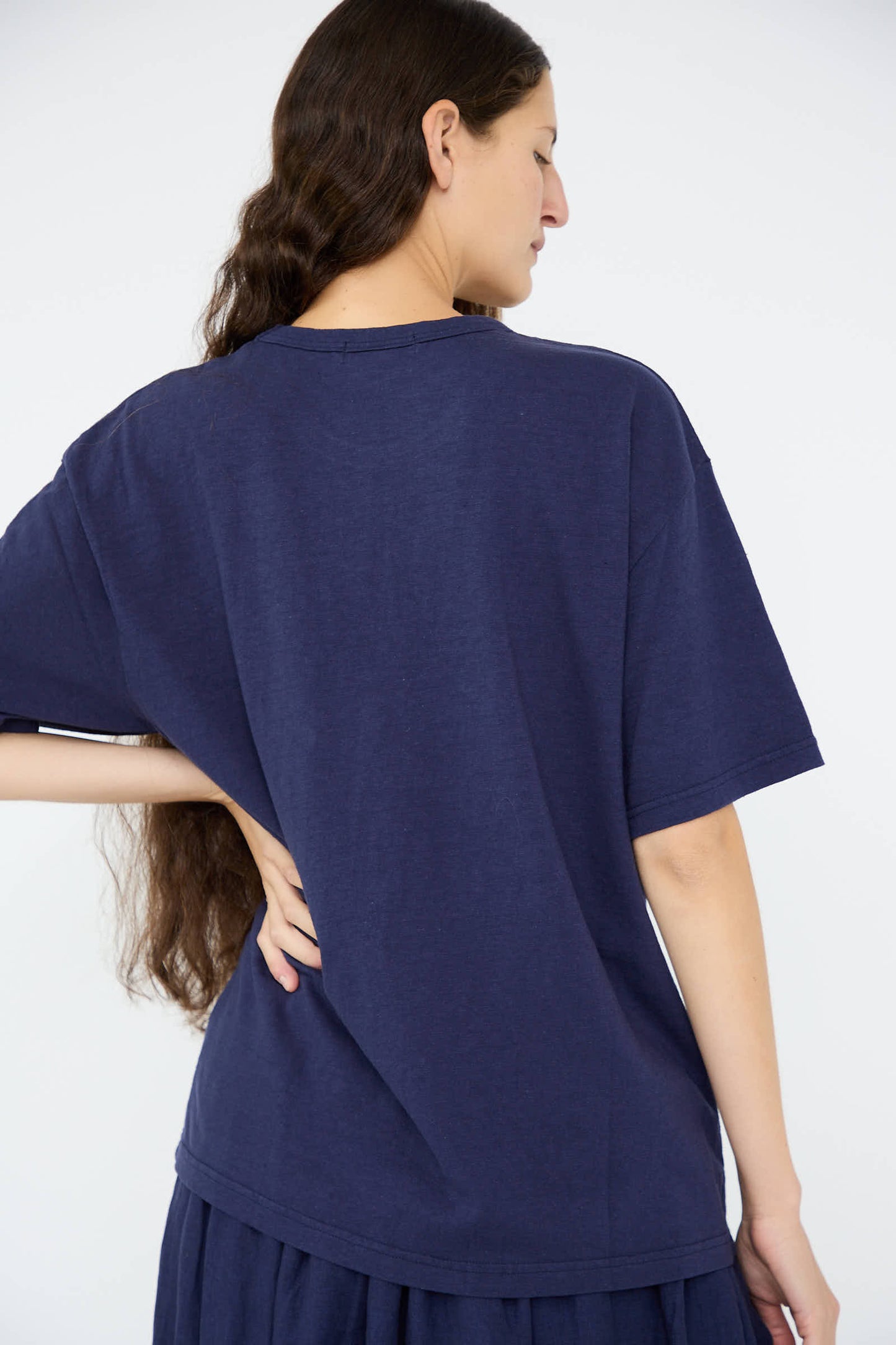 A person with long hair stands facing away, wearing a Ichi Antiquités Cotton T-Shirt in navy, crafted from medium-weight cotton, with their left hand on their hip.