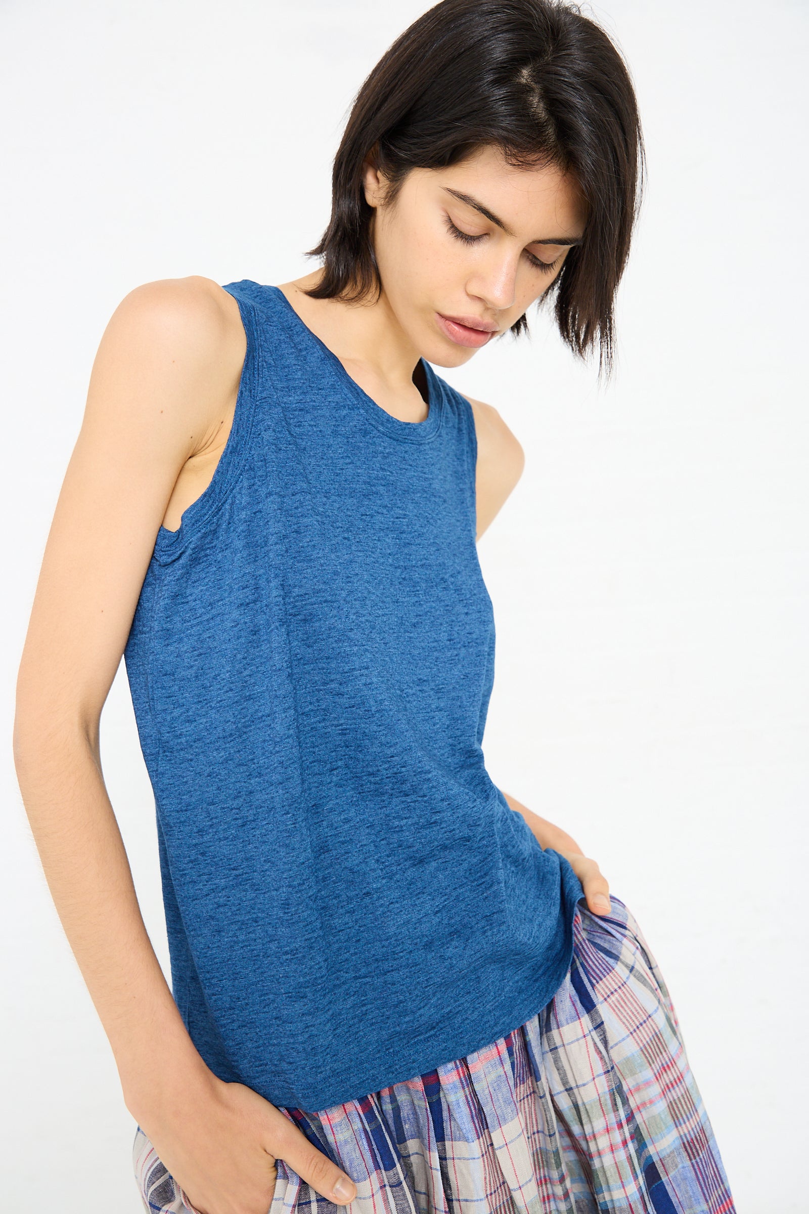 A person with short dark hair wearing an Ichi Antiquités Cotton Tank in Blue and a plaid skirt looks down while posing against a white background.