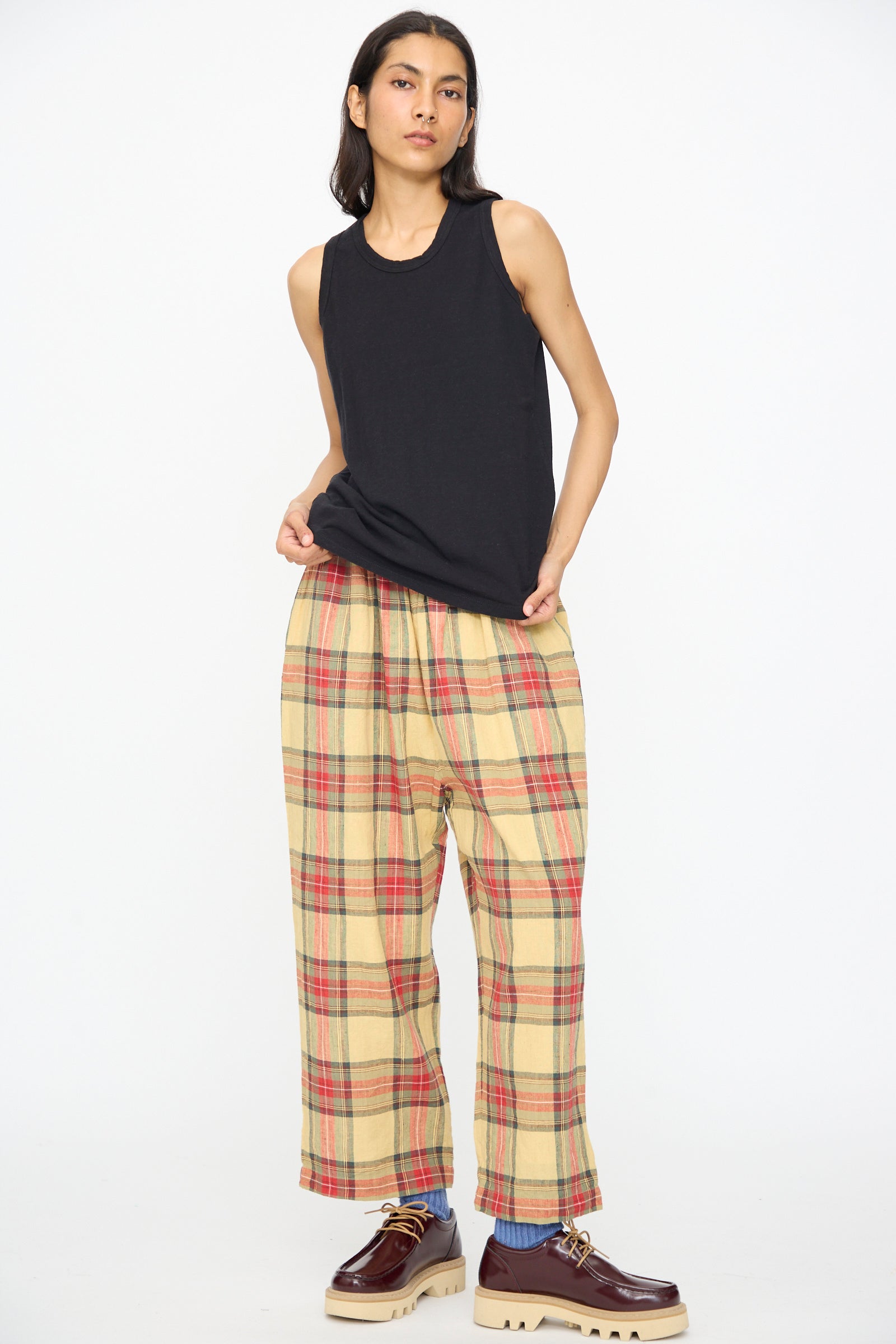 A person stands against a white background wearing the Ichi Antiquités Cotton Tank Top in Black, loose beige plaid pants with red checks, and brown thick-soled shoes.
