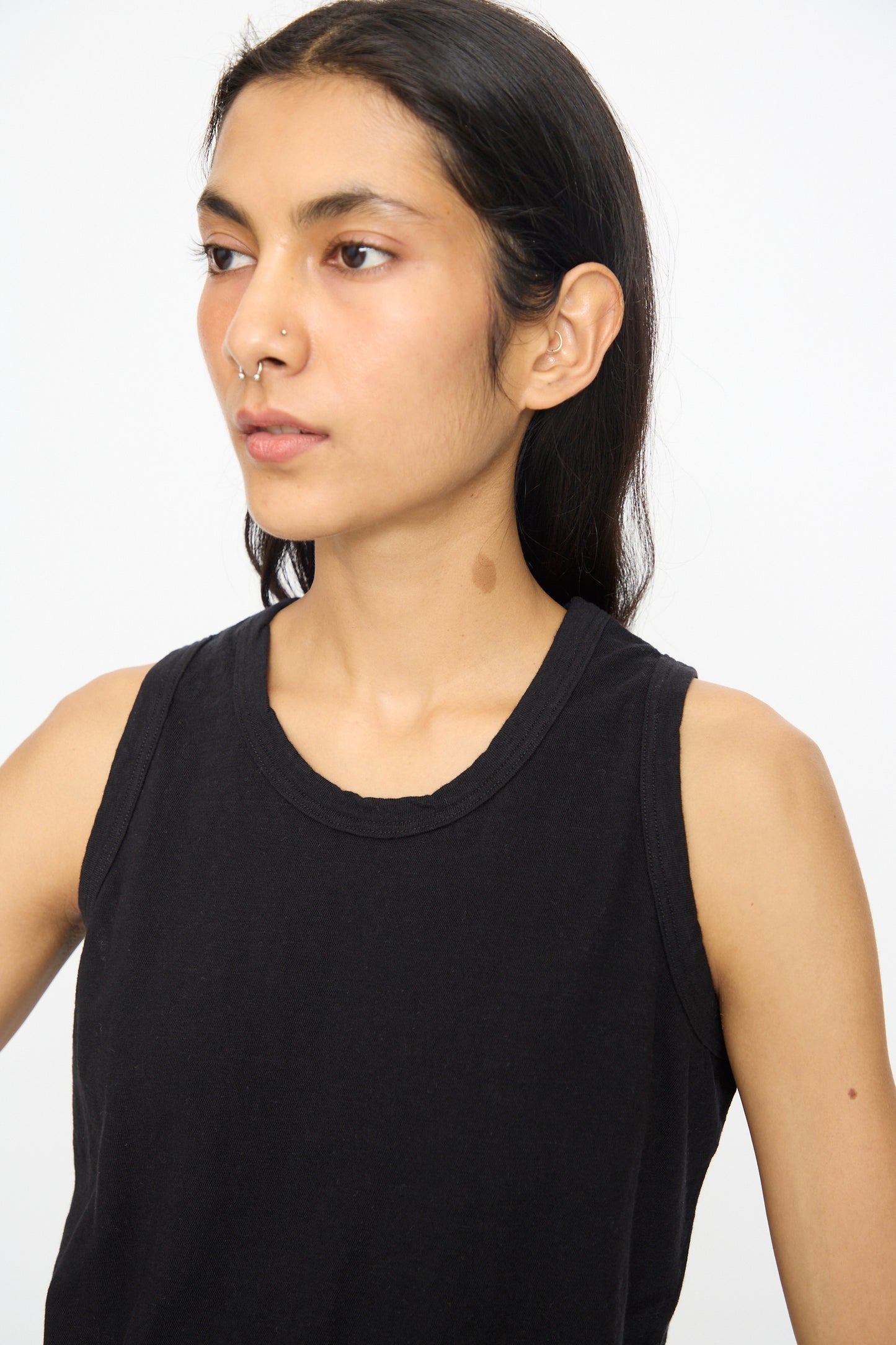 A person with long dark hair wears the Cotton Tank Top in Black by Ichi Antiquités, paired with silver nose jewelry and featuring a visible neck birthmark, standing before a plain light background.