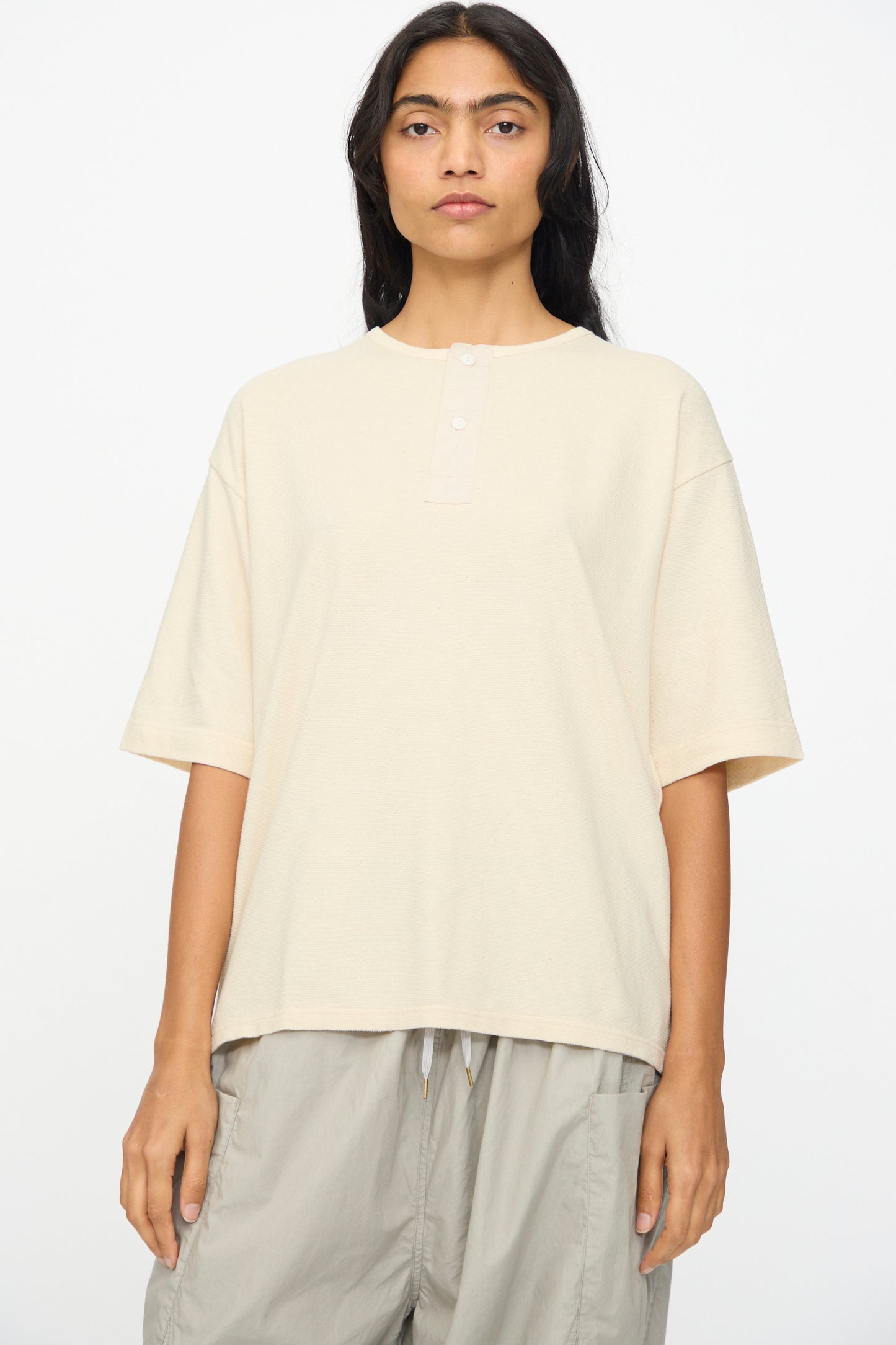 A person with long dark hair wears the Ichi Antiquités Cotton Thermal Henley Neck Pullover in Ivory, paired with light gray drawstring pants, standing against a plain white background.