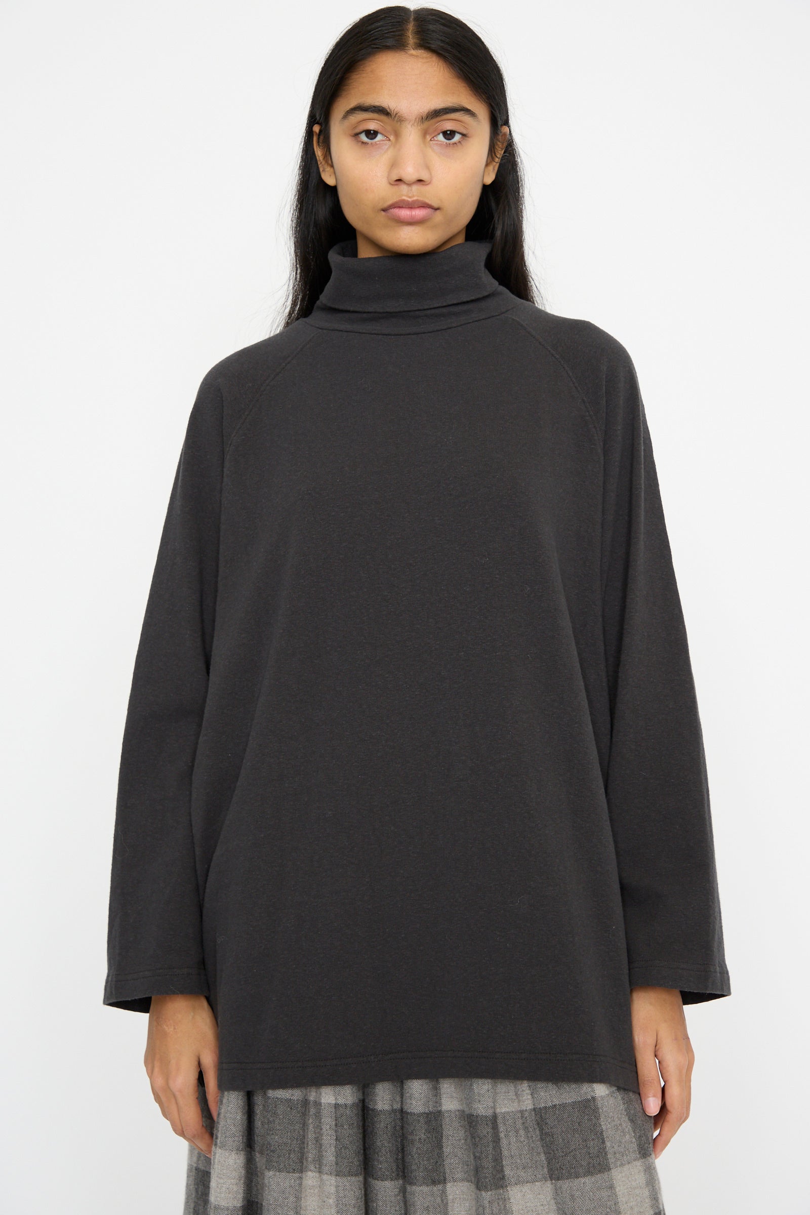 A model dressed in an oversized Ichi Antiquités Cotton Turtleneck in Black and a gray plaid skirt is posed against a simple background.