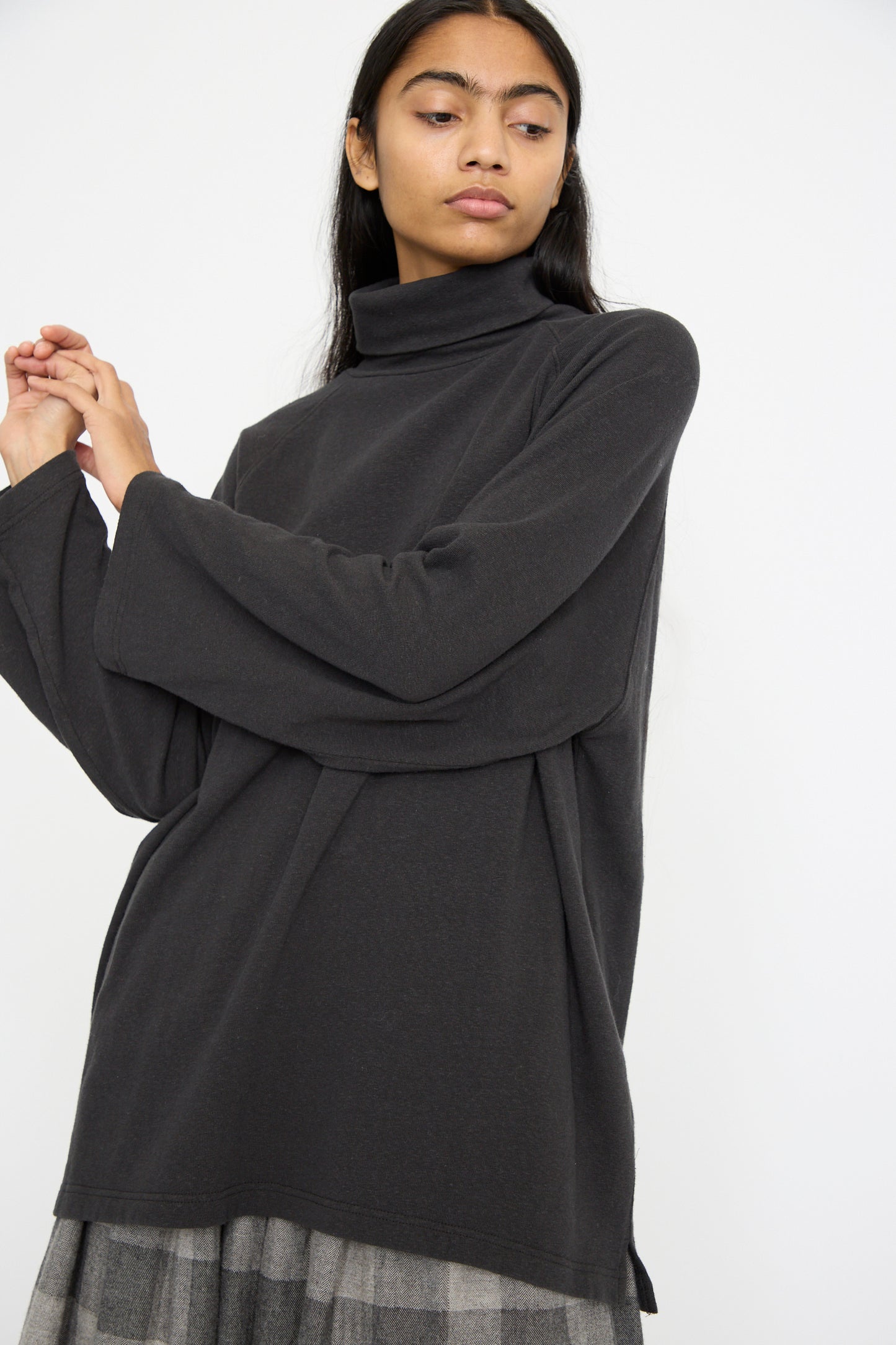 A person wears a black cotton turtleneck by Ichi Antiquités with an oversized fit and a gray plaid skirt, standing against a plain white background.