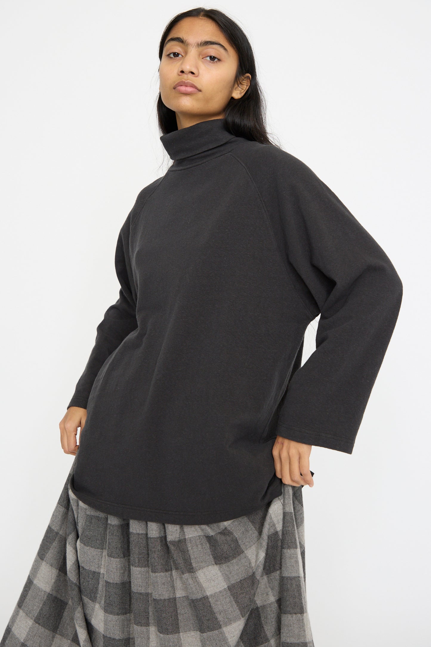 A person stands against a pristine white backdrop in an oversized Ichi Antiquités Cotton Turtleneck in Black paired with a gray checkered skirt.