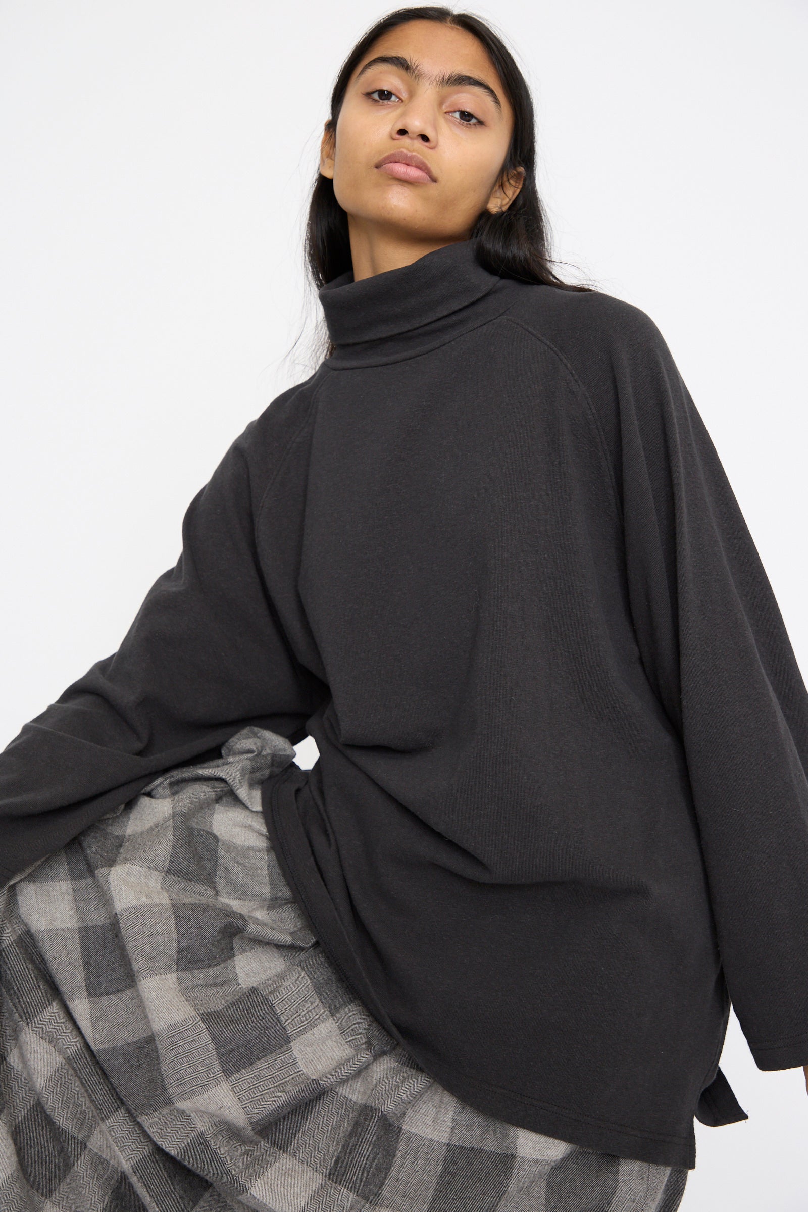 A person with long dark hair wears an oversized Cotton Turtleneck in Black by Ichi Antiquités, paired with a gray checkered skirt, against a plain white background.