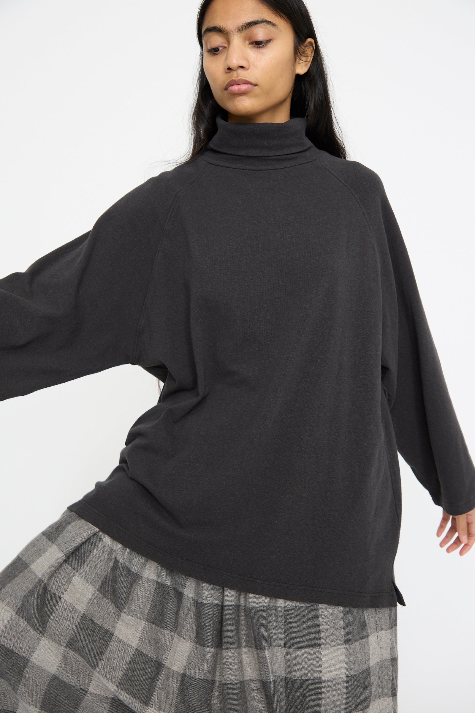 Wearing an Ichi Antiquités Cotton Turtleneck in Black and a plaid skirt, a person poses against a plain background and looks down.