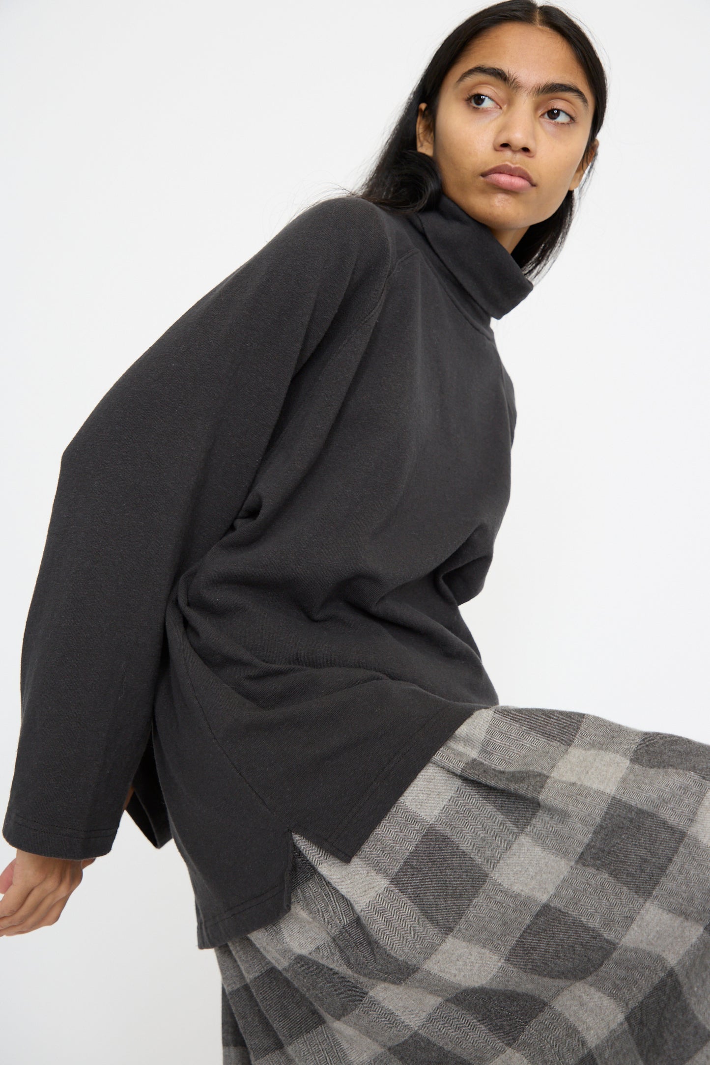 A person poses against a white background, wearing Ichi Antiquités' oversized-fit Cotton Turtleneck in Black paired with a plaid skirt.