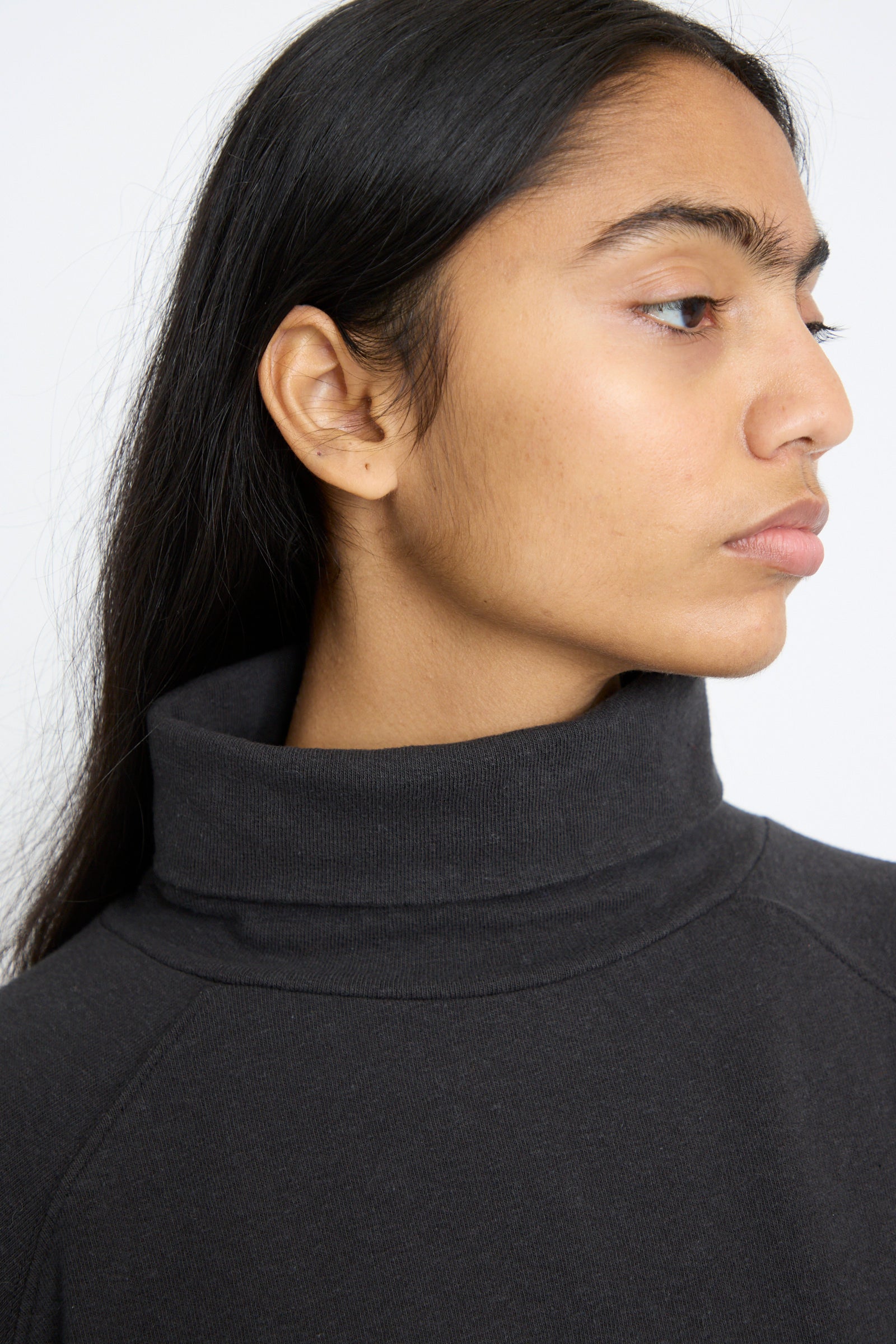 A person with long dark hair wears an oversized Cotton Turtleneck in Black, by Ichi Antiquités, and looks to the side against a plain background.