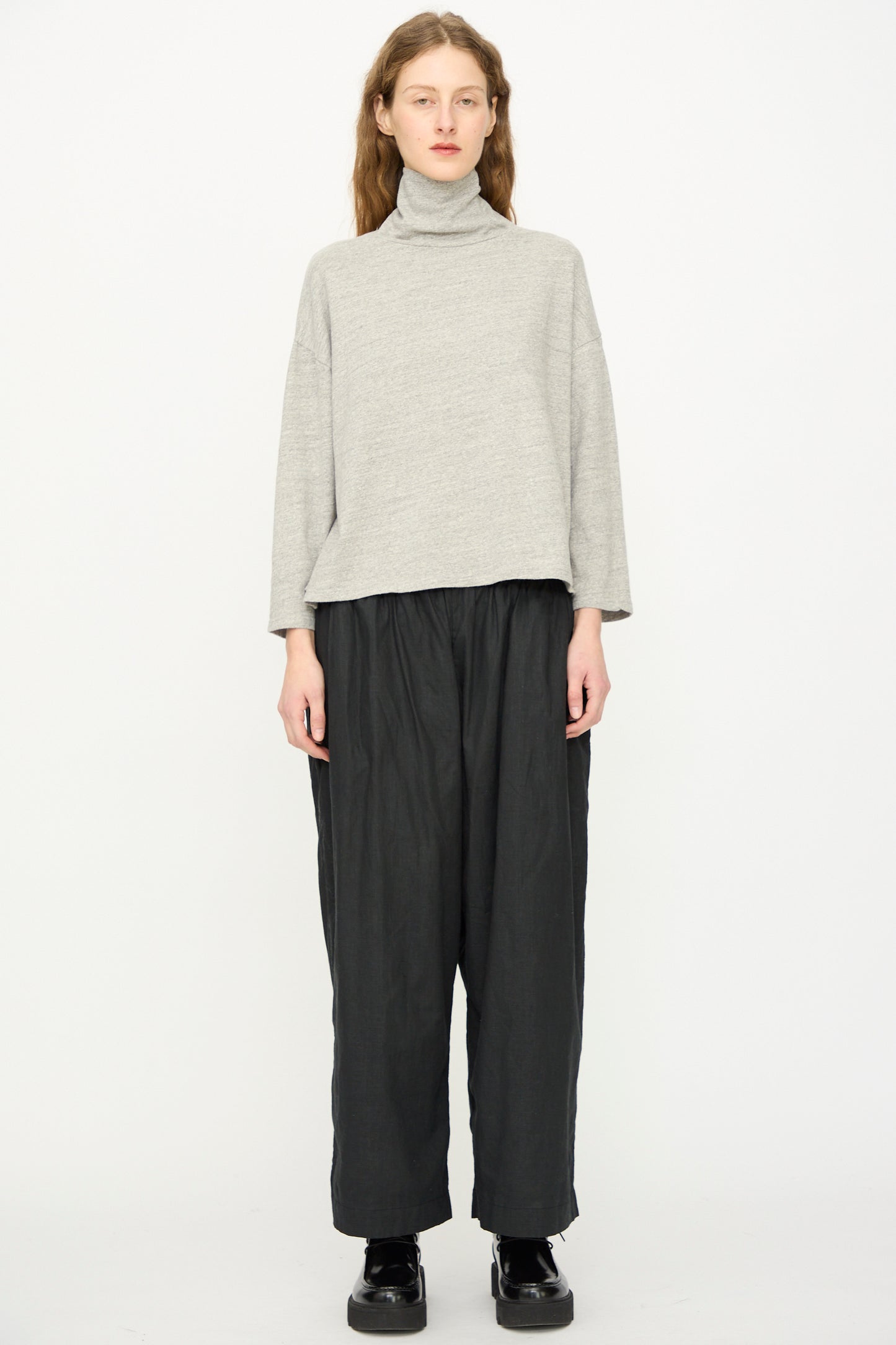 A person stands against a white background wearing the Ichi Antiquités Cotton Turtleneck in Gray, along with loose black pants and black shoes.