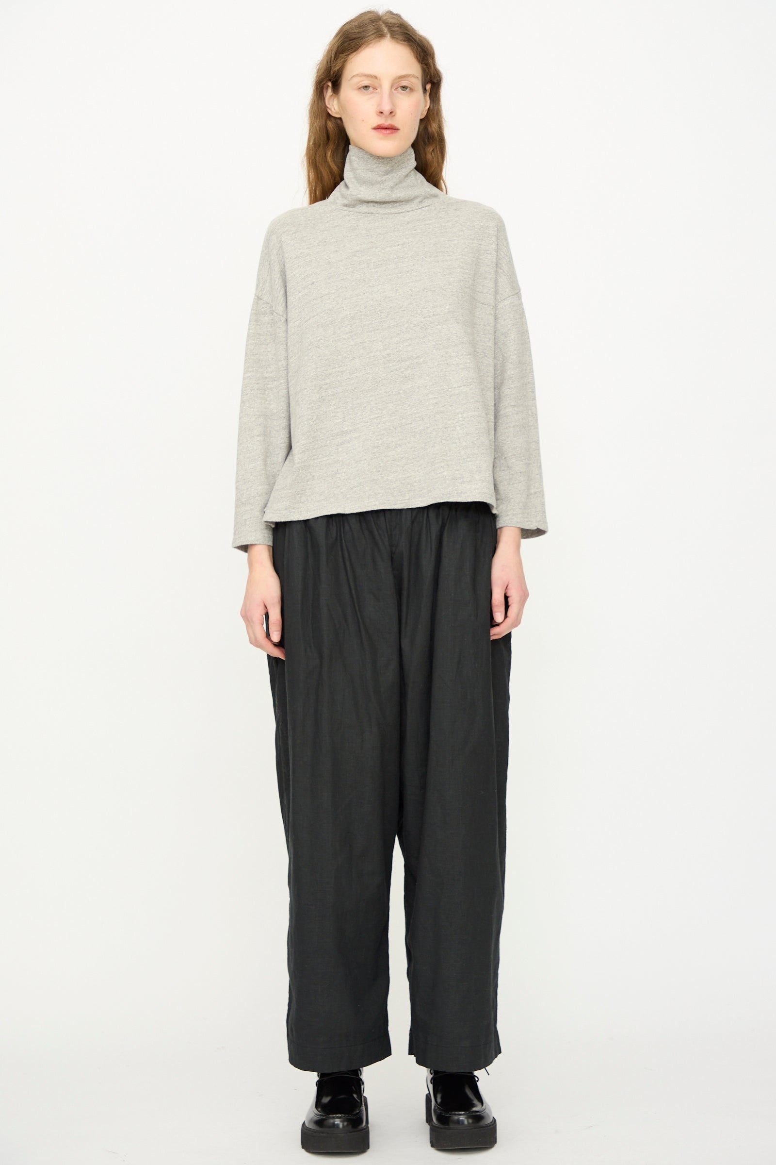 A person stands against a white background wearing the Ichi Antiquités Cotton Turtleneck in Gray, along with loose black pants and black shoes.