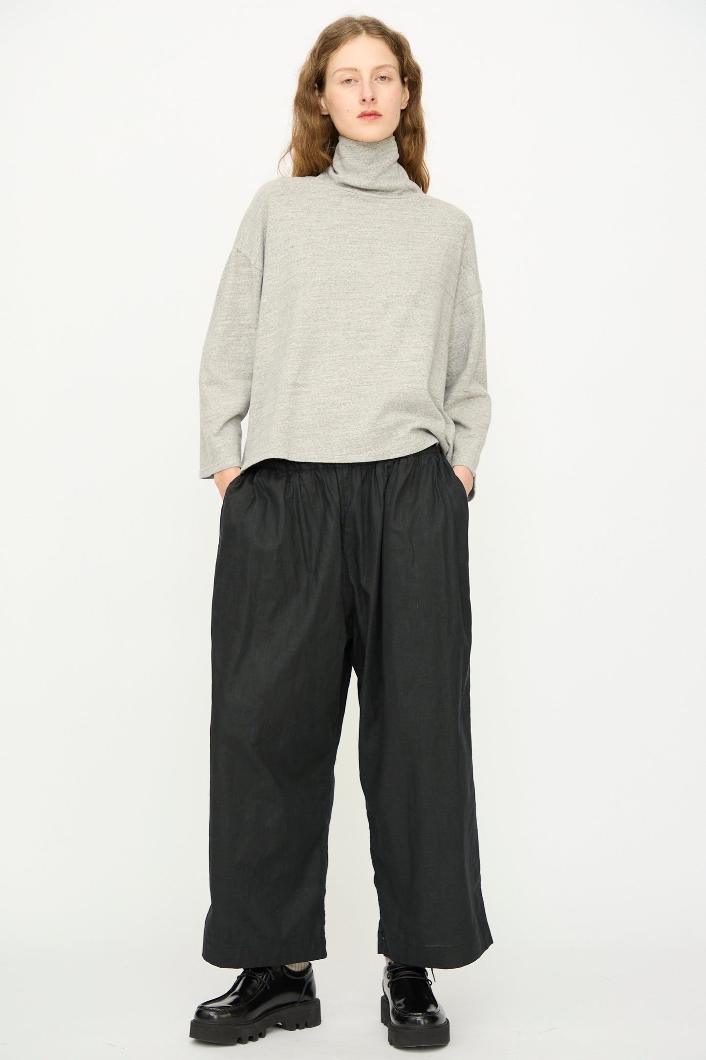 A person stands against a plain background wearing the Ichi Antiquités Cotton Turtleneck in Gray, wide black pants, and black shoes, with hands in pockets.